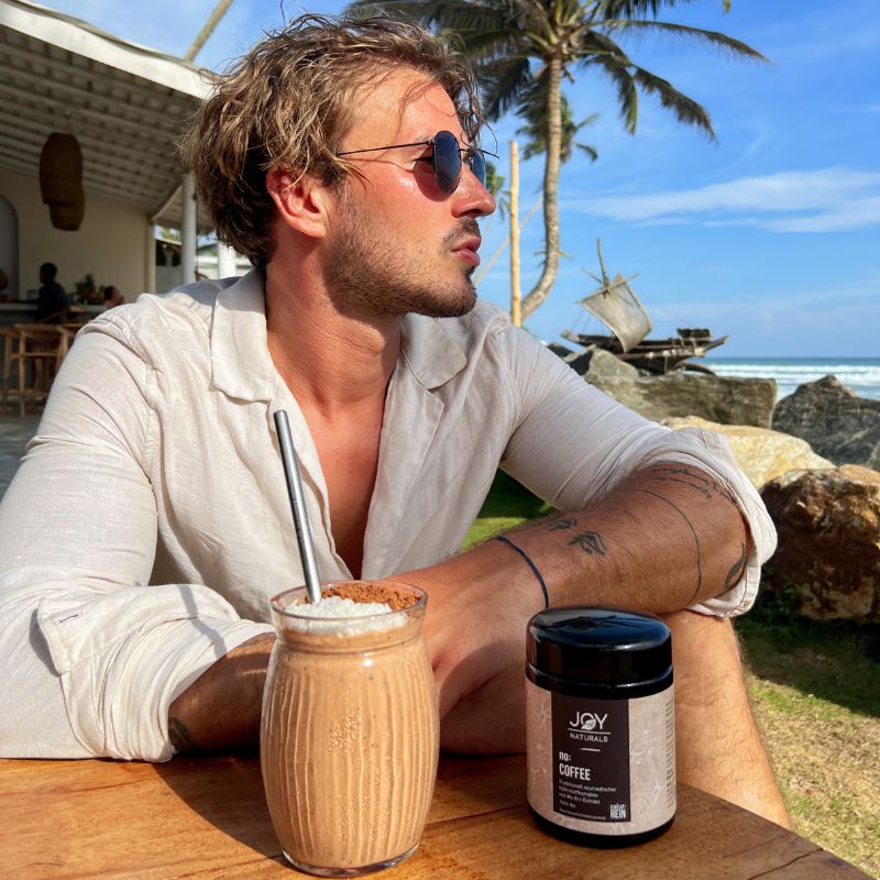 Mann mit Sonnenbrille sitzt am Tisch. Neben ihm ein Glasgetränk mit Strohhalm und ein schwarzes Glas mit Aufschrift 'JOY NATURALS no: COFFEE'.