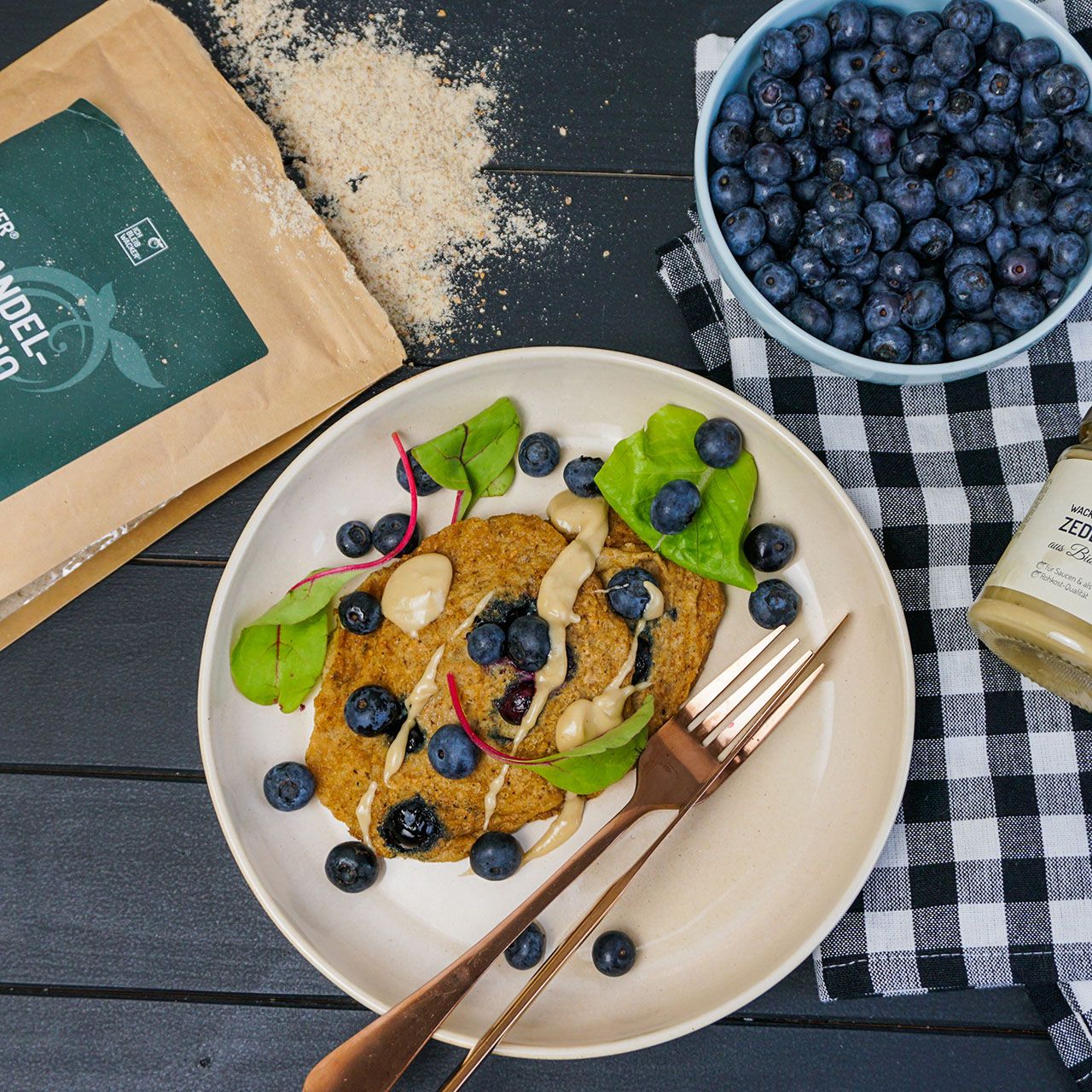 Teller mit Pfannkuchen, Blaubeeren und Sauce. Daneben eine Tüte mit Erdmandelflocken und eine Schüssel mit Blaubeeren.