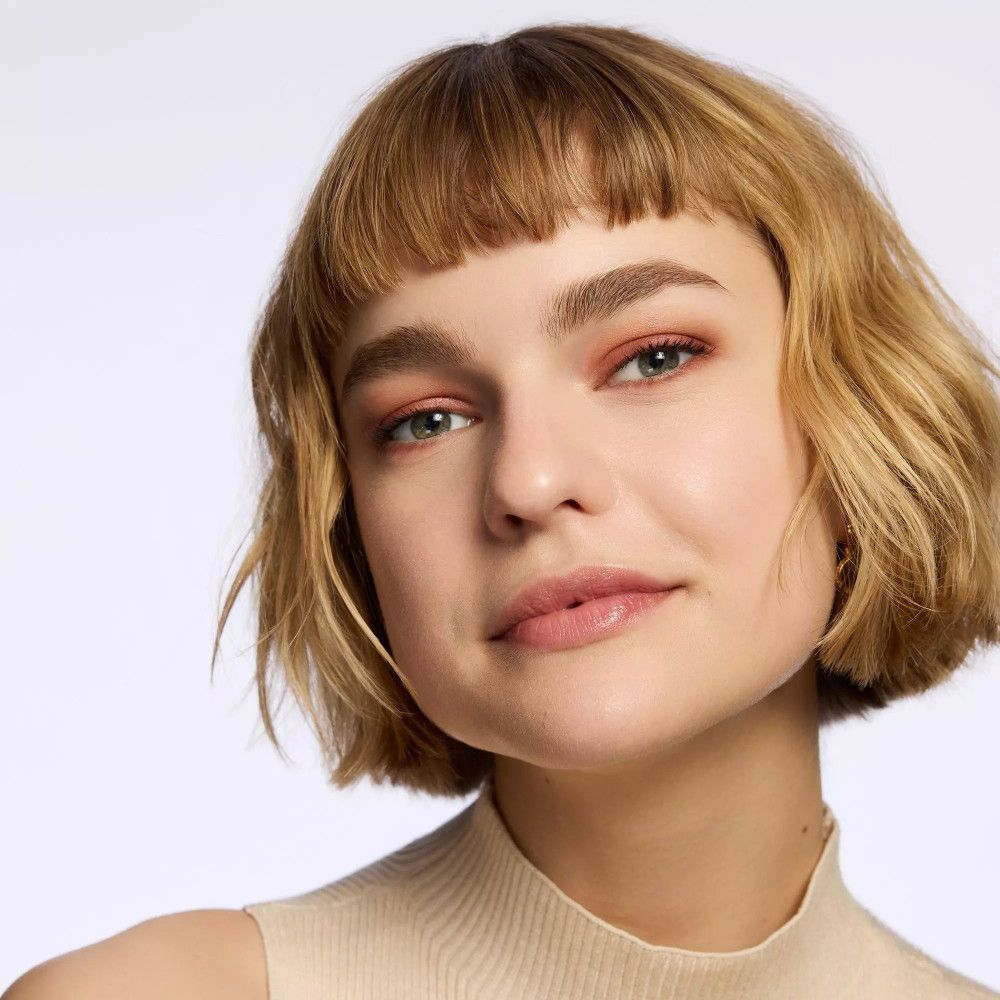 Porträt einer Frau mit geschminkten Augen und Wangen. Rouge und Lidschatten in warmen Tönen. Kurze blonde Haare.