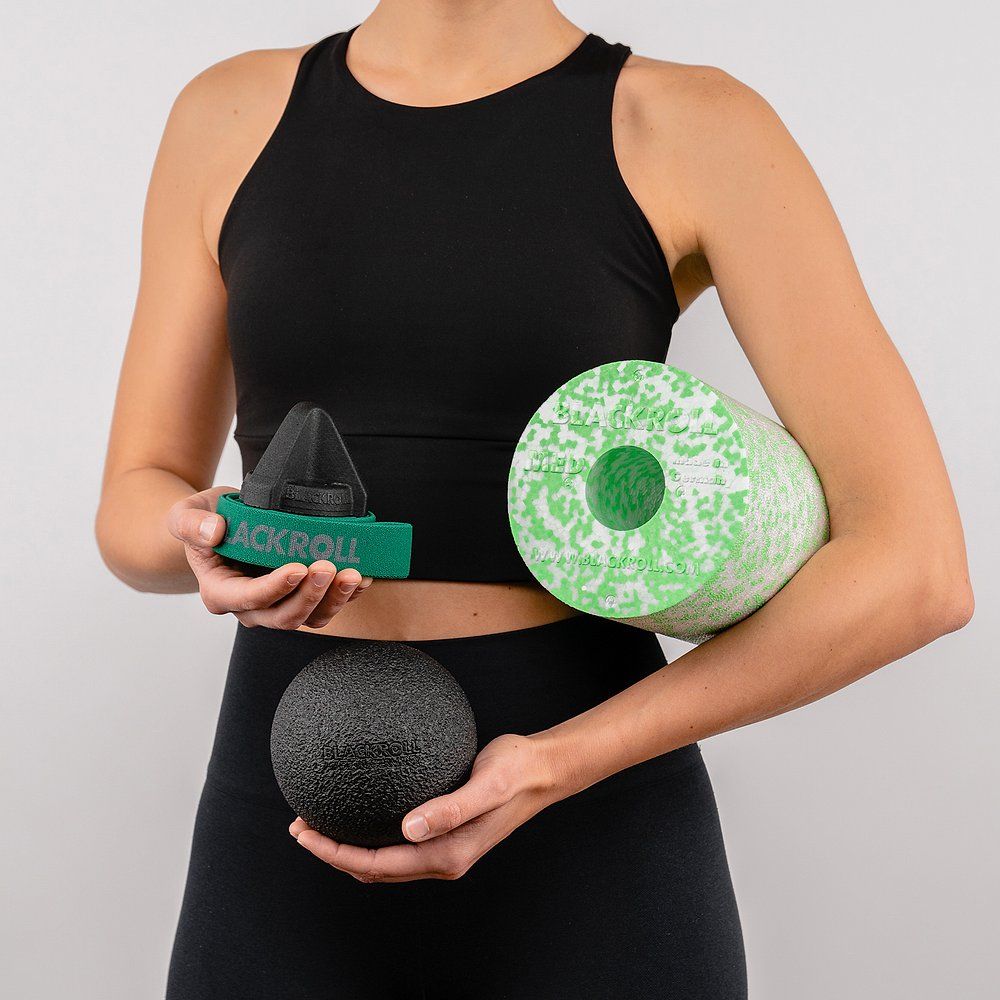 Person hält drei Produkte: schwarze Rolle, grünes Dreieck, grüne Rolle. Sportkleidung.