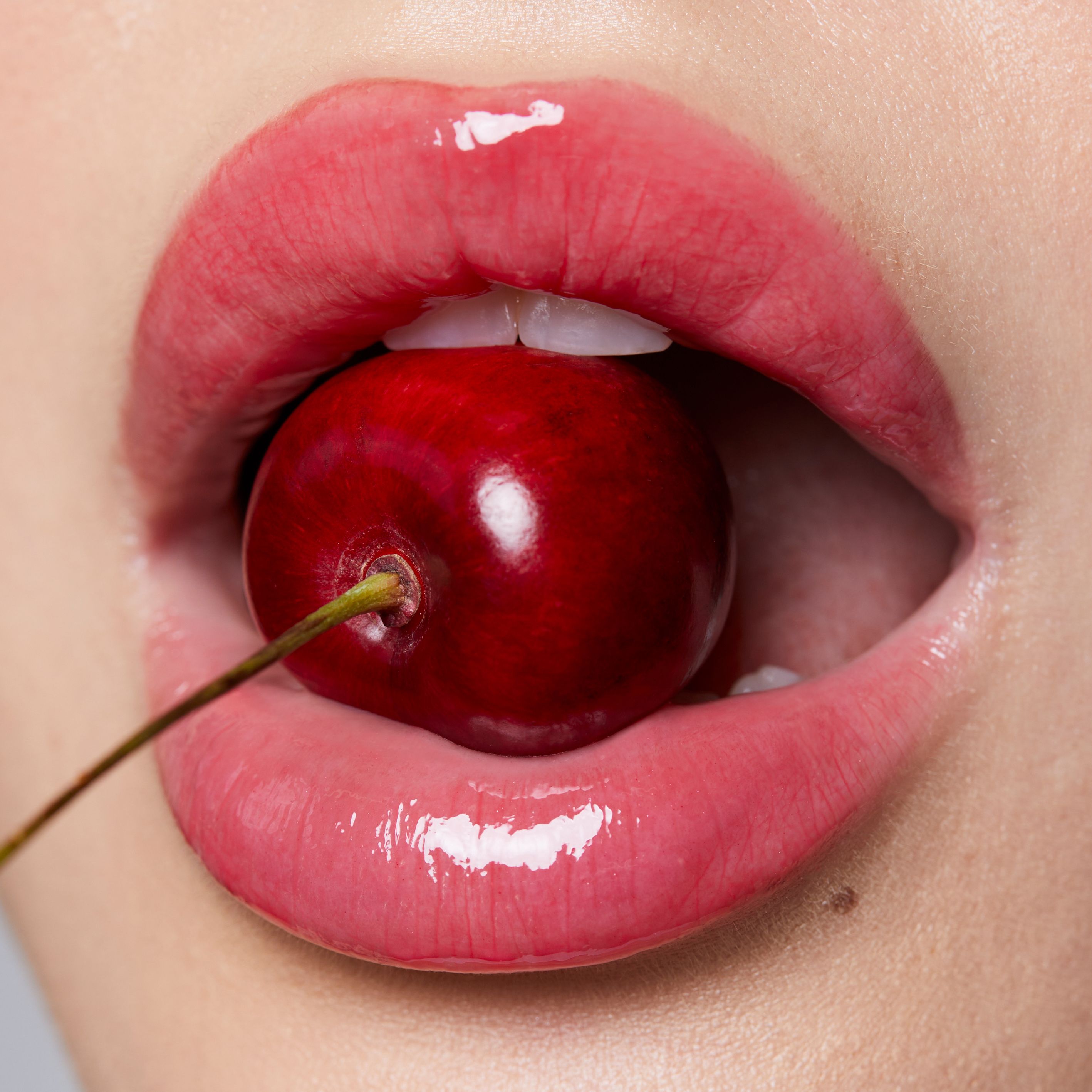 Nahaufnahme von Lippen mit rotem Lipgloss. Eine Kirsche wird in den Mund gehalten.