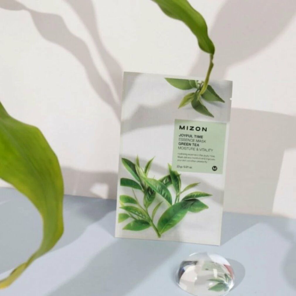 Produktverpackung mit grünen Blättern. Text: MIZON Joyful Time Essence Mask Green Tea. Auf hellem Untergrund mit Dekoration.