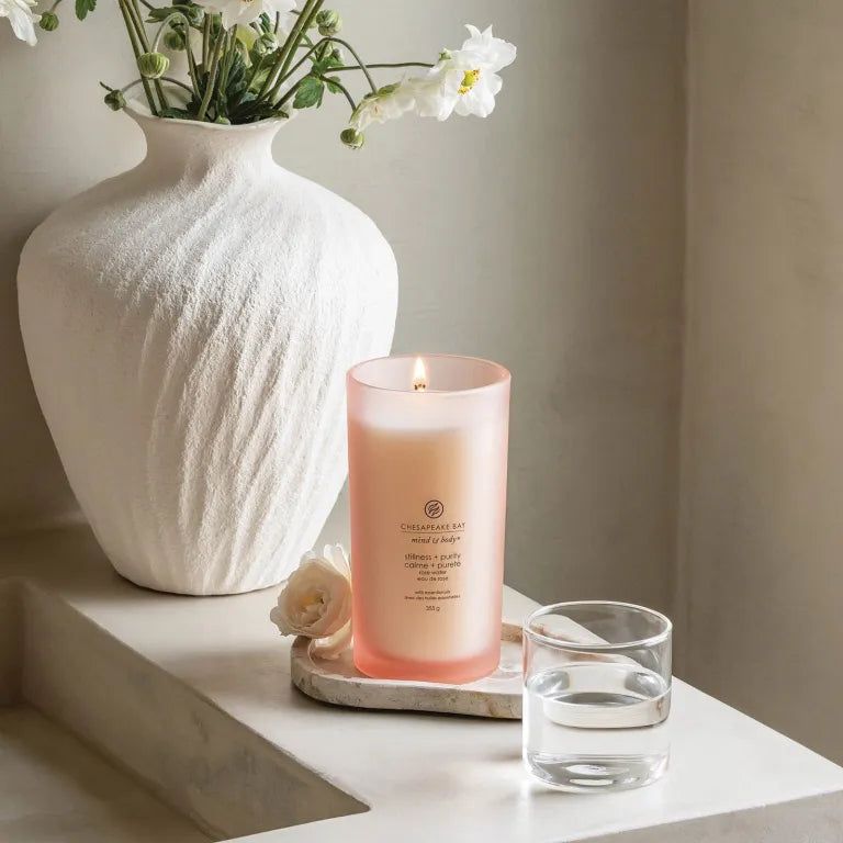 Brennende rosa Kerze. Aufschrift: Chesapeake Bay, mind & body, stillness + purity, calme + pureté, rose water, 355g. Neben Glas Wasser.