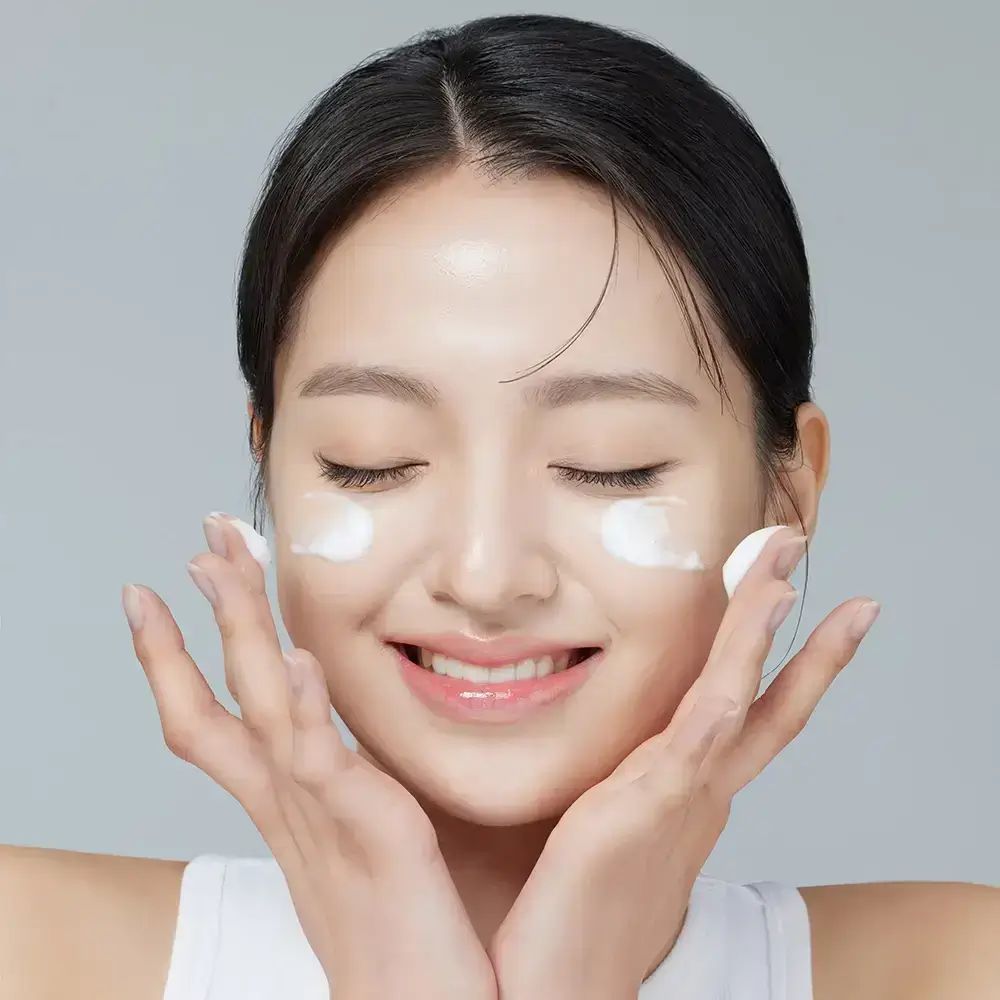 Frau mit geschlossenen Augen, trägt Creme auf. Hände am Gesicht. Weißes Top.