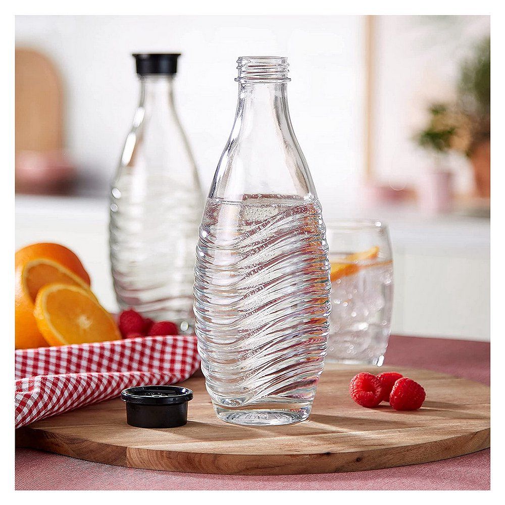 Eine SodaStream Glasflasche mit schwarzem Deckel, gefüllt mit sprudelndem Wasser, auf einem Holzbrett mit Früchten und einem Glas Wasser.