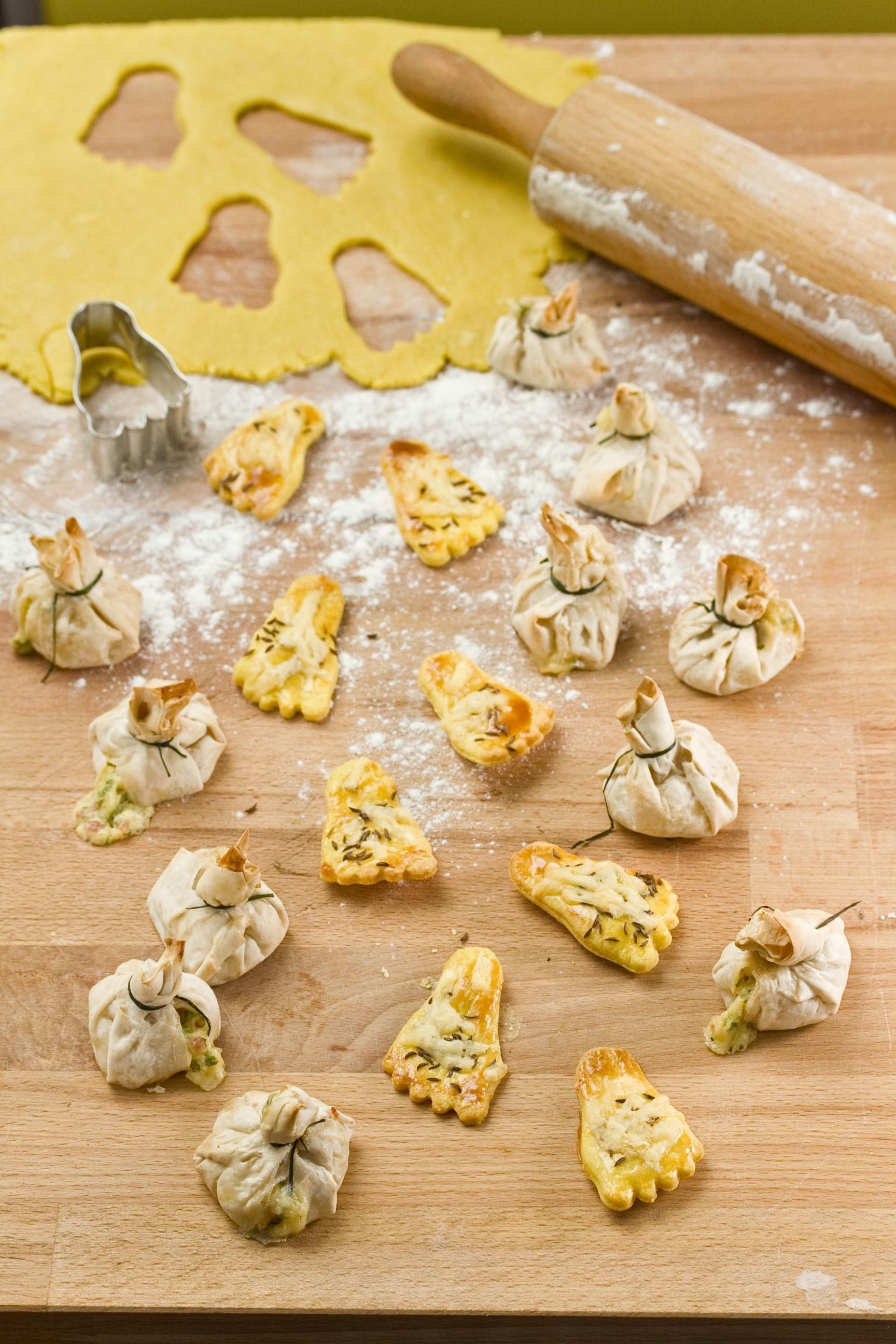 Holzbrett mit Teig, Ausstecher, Nudeln und gefüllten Teigtaschen. Einige sind gefaltet, andere gebacken, mit Kräutern.