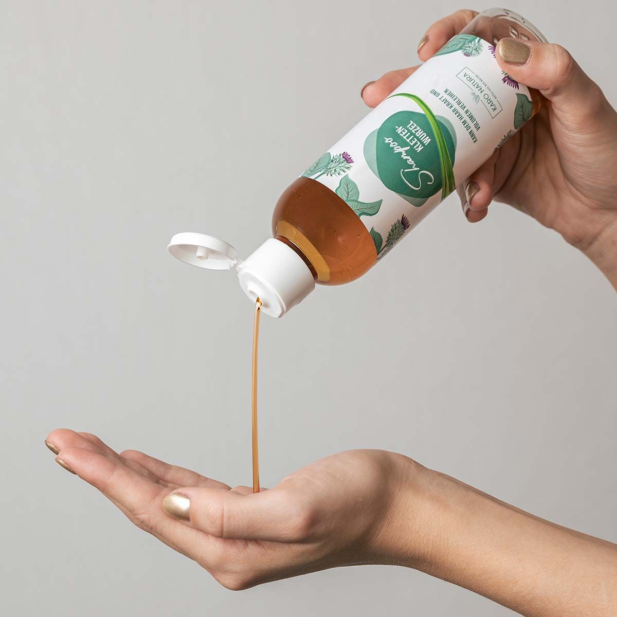 Eine Flasche mit Shampoo, aus der Flüssigkeit in eine Hand gegossen wird. Weißer Deckel. Etikett mit Schriftzug Shampoo Klettenwurzel. Karo Natura Logo.