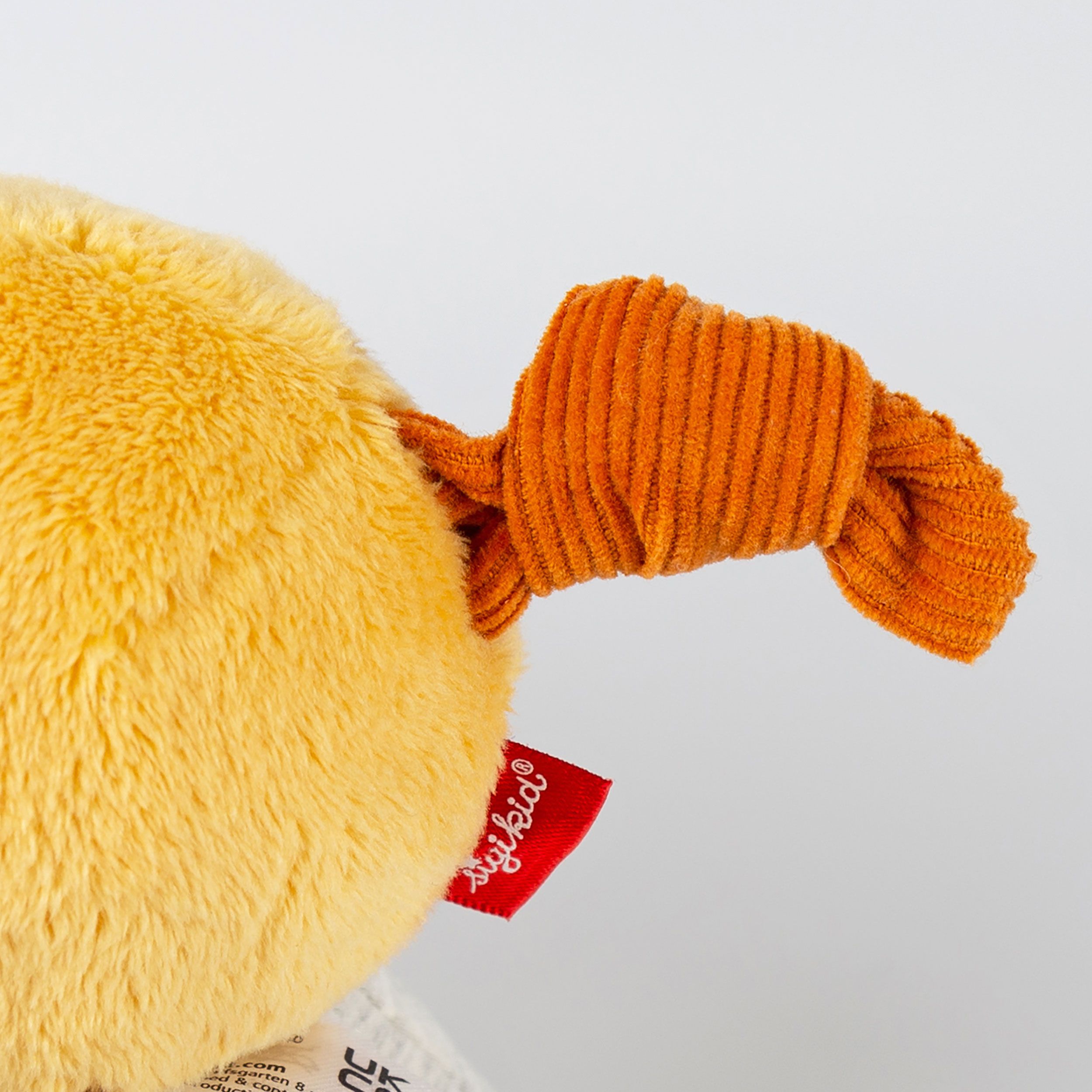 Nahaufnahme des orangefarbenen Ohres und des Etiketts einer gelben Stoffgiraffe. Rotes Etikett mit Logo.