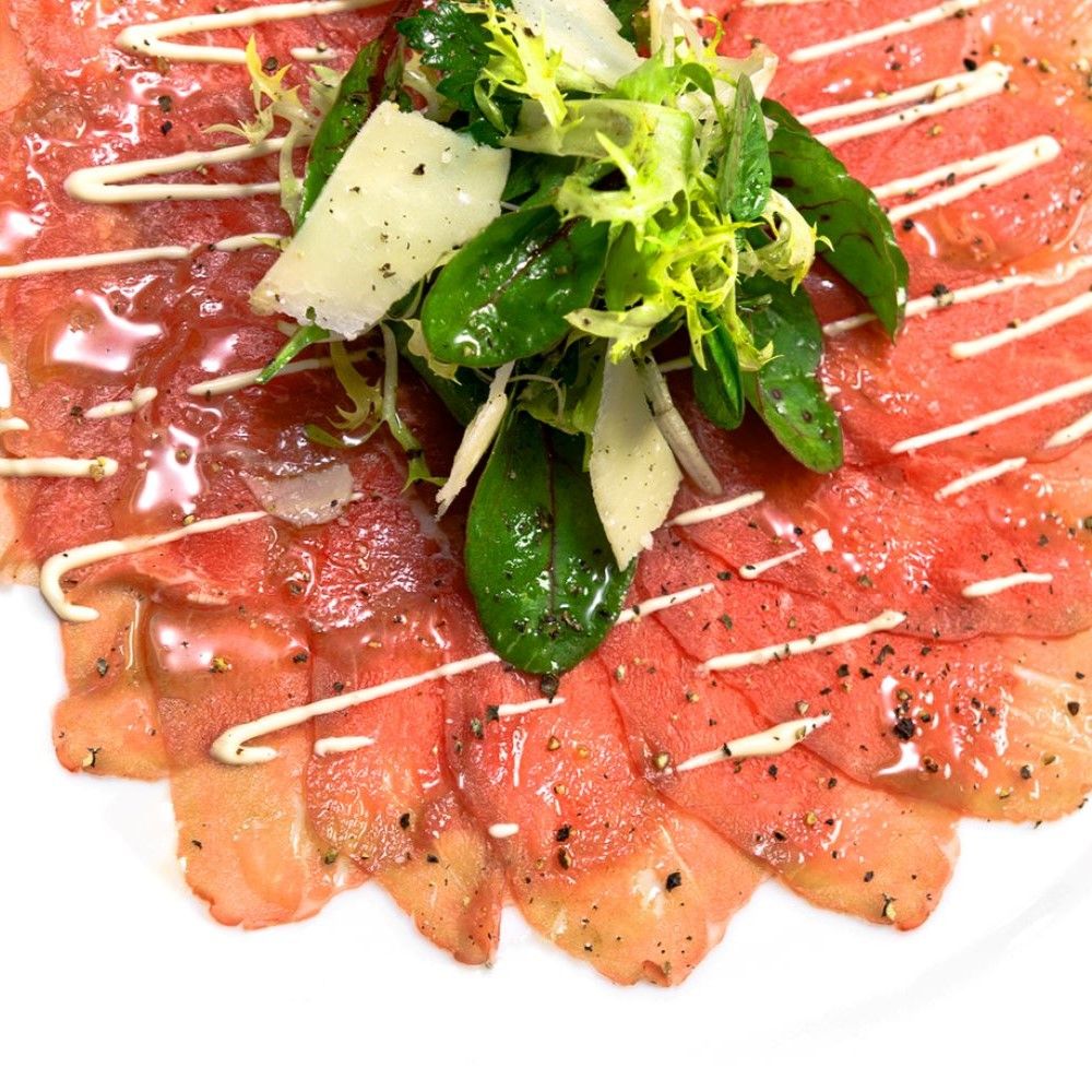 Carpaccio mit Salat und Parmesan. Dünne, rote Fleischscheiben, mit weißer Sauce beträufelt, mit Salat und Parmesan.