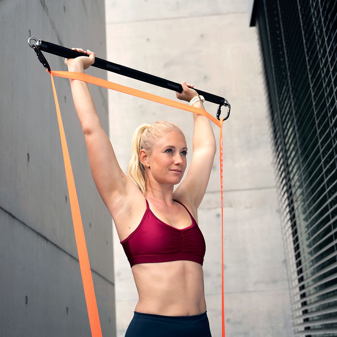 Frau mit Fitnessband und Stange. Sie steht vor einer Betonwand. Das Band ist orange.