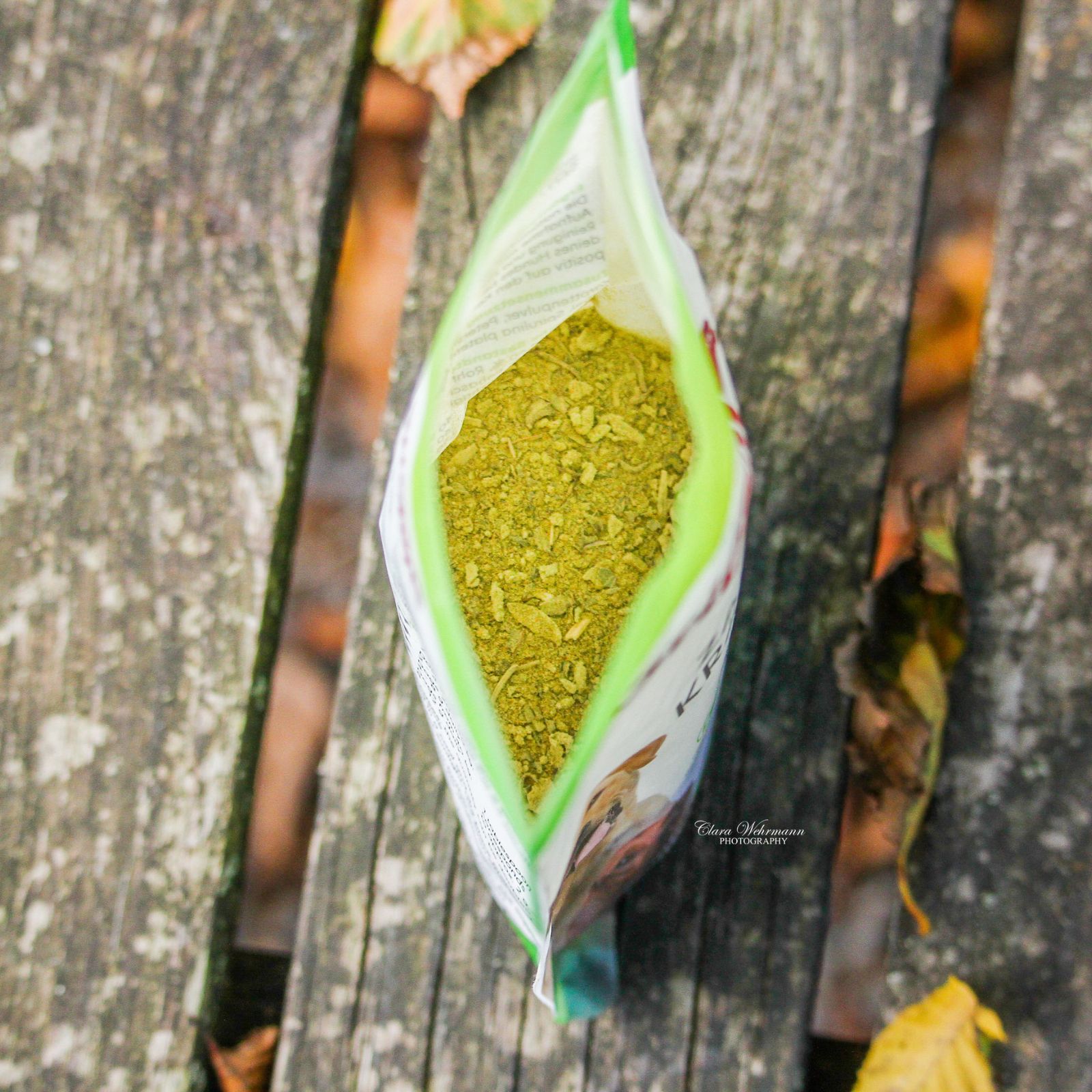 Geöffnete grüne Tüte mit Stiefel Zahnkräuter. Inhalt: getrocknete Kräuter. Auf Holzuntergrund. Sichtbar: Kräuter, Verpackung, Text.