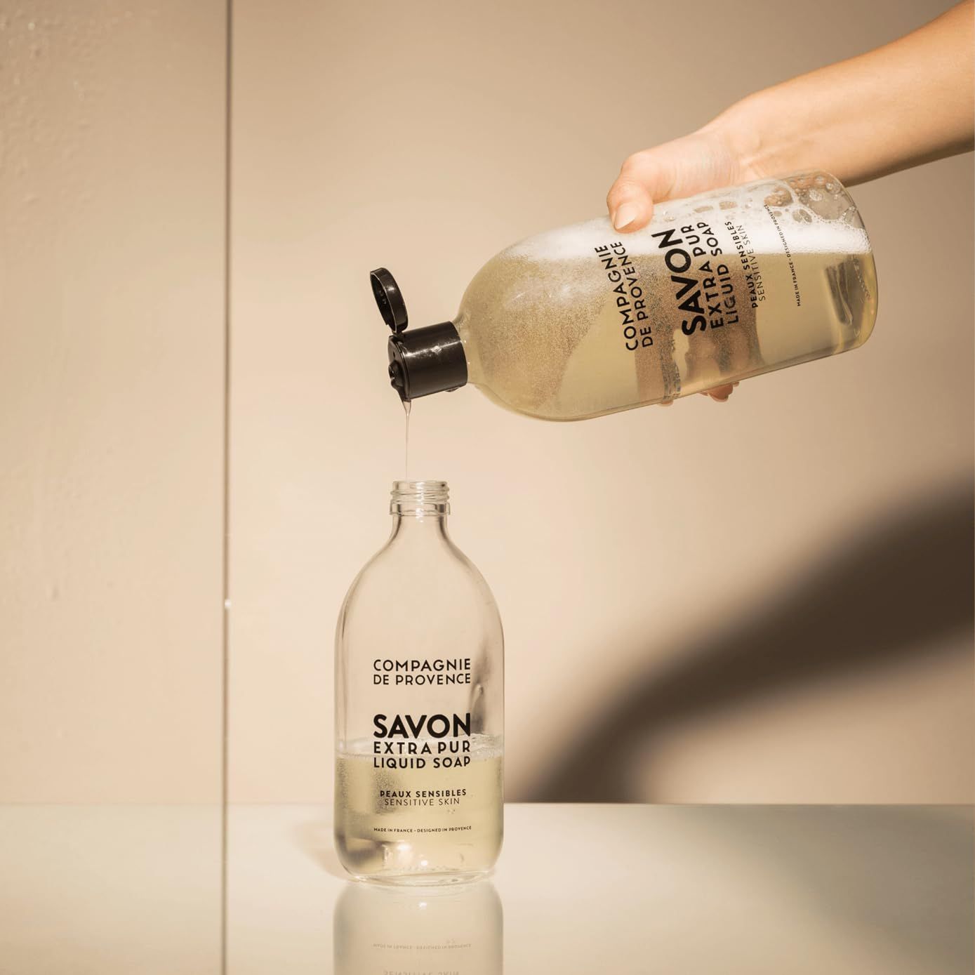 Hand gießt Flüssigkeit aus einer Flasche in eine andere. Text: Compagnie de Provence, Savon Extra Pur Liquid Soap.