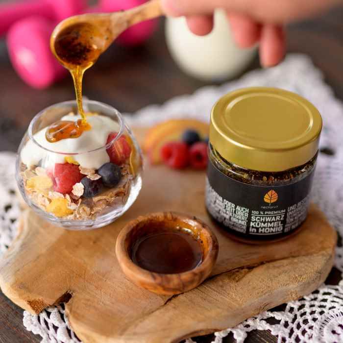 Honig wird mit einem Löffel in ein Glas mit Joghurt und Früchten gegossen. Honigglas mit goldenem Deckel daneben.