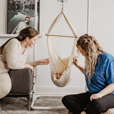 Zwei Frauen betrachten ein Baby in einer Babyhängematte. Beige Stoff, Holzstab, Metallgestell.