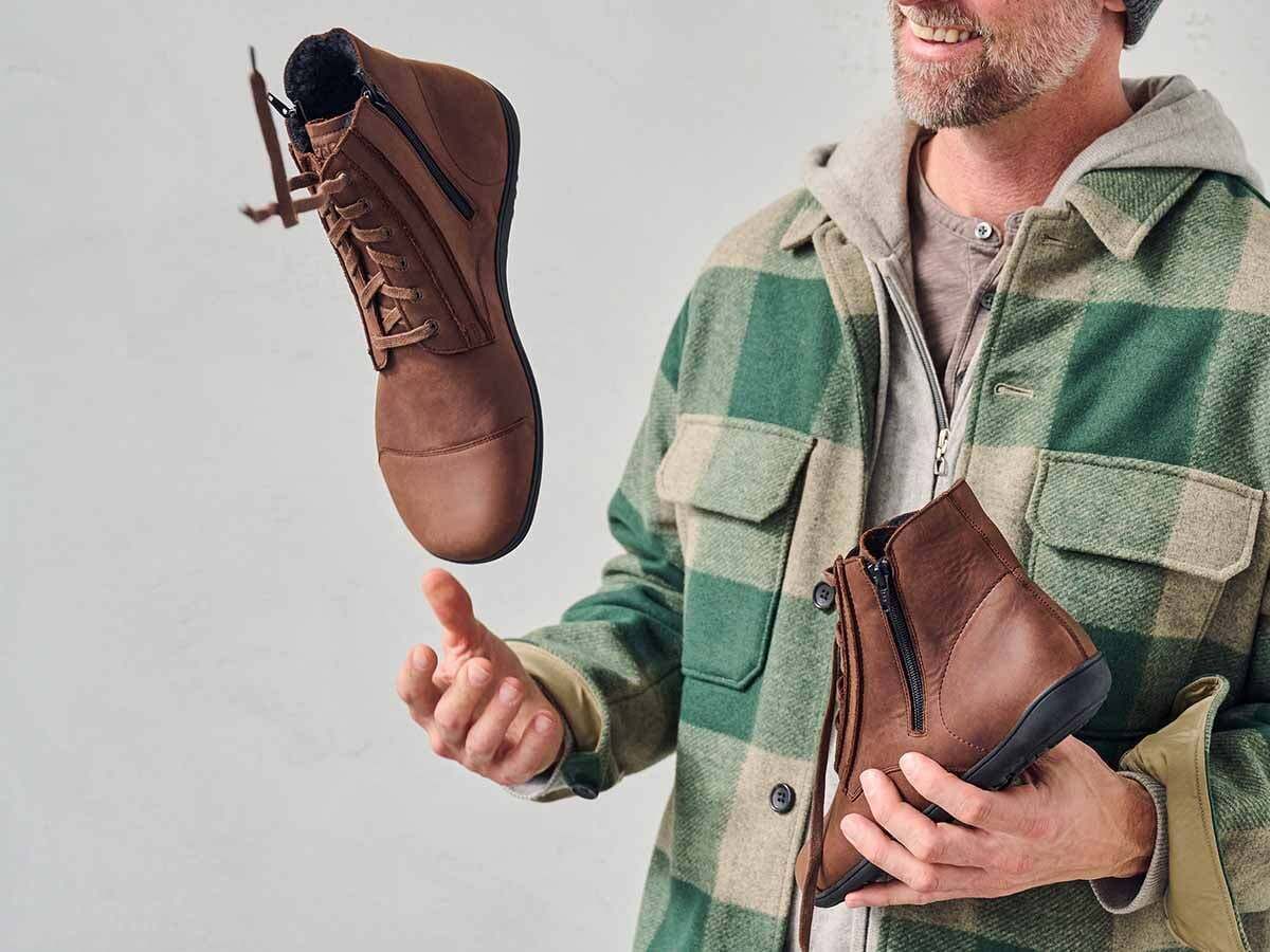 Mann hält zwei braune Stiefeletten. Schnürung und Reißverschluss. Breite Passform. Lederoptik. Schwarze Sohle. Eine schwebt.