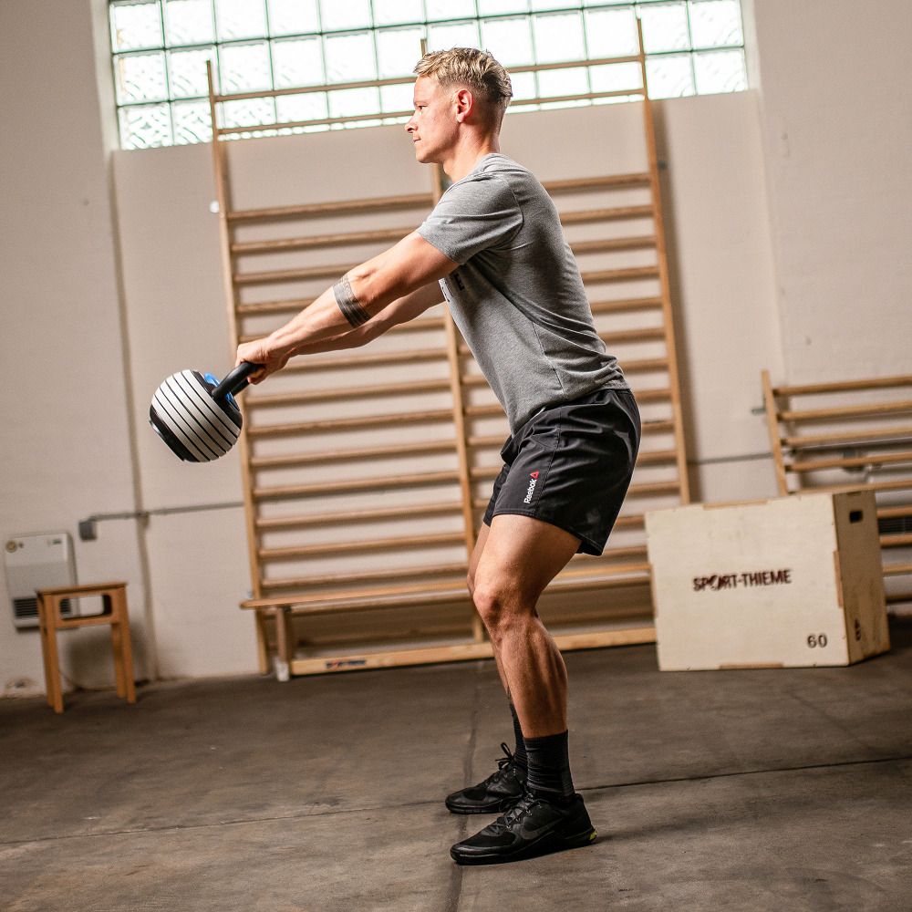 Kettlebell Vario, schwarz-weiß, in Gebrauch. Athlet in Bewegung, hält das Gerät. Hintergrund: Fitnessraum.