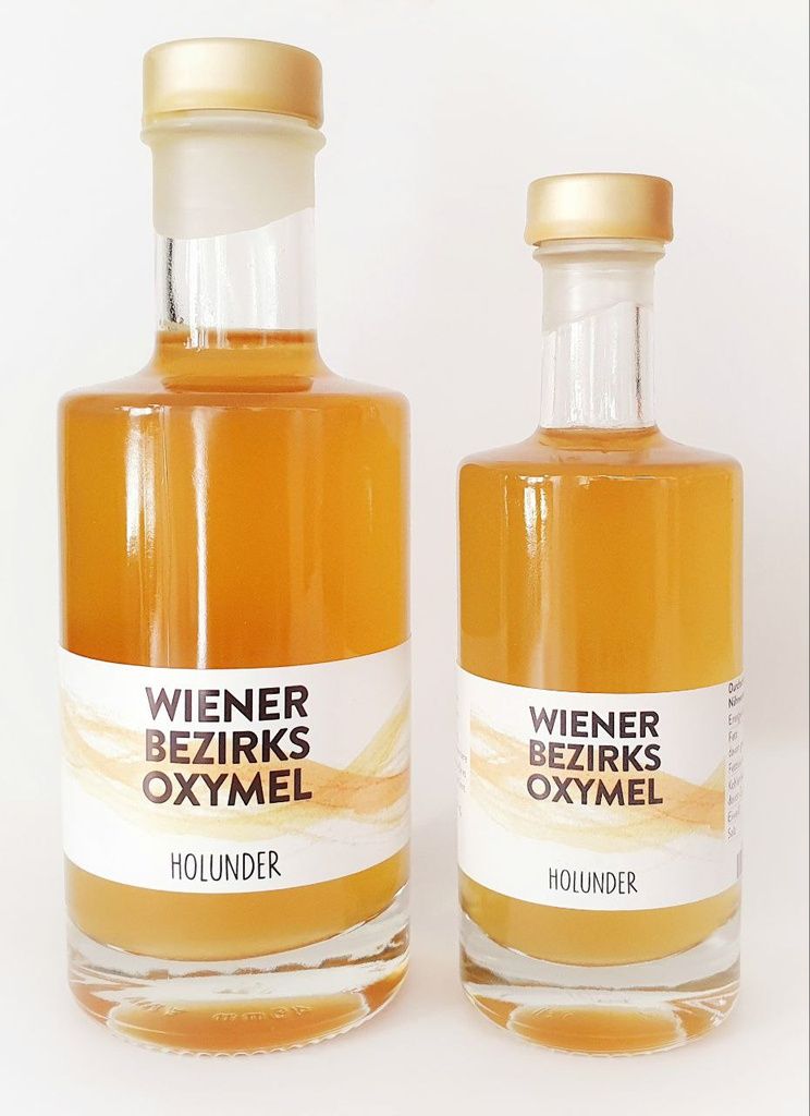 Zwei Glasflaschen mit goldfarbenem Inhalt. Etikett mit Text: Wiener Bezirks Oxymel Holunder. Goldfarbene Verschlüsse.