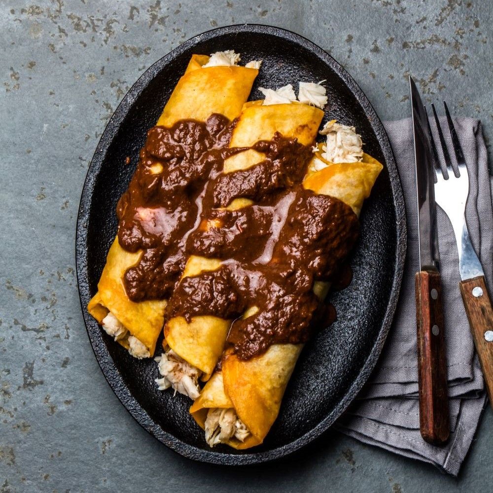 Enchiladas auf schwarzer Platte. Mit brauner Soße bedeckt. Neben Gabel und Messer auf grauem Untergrund.