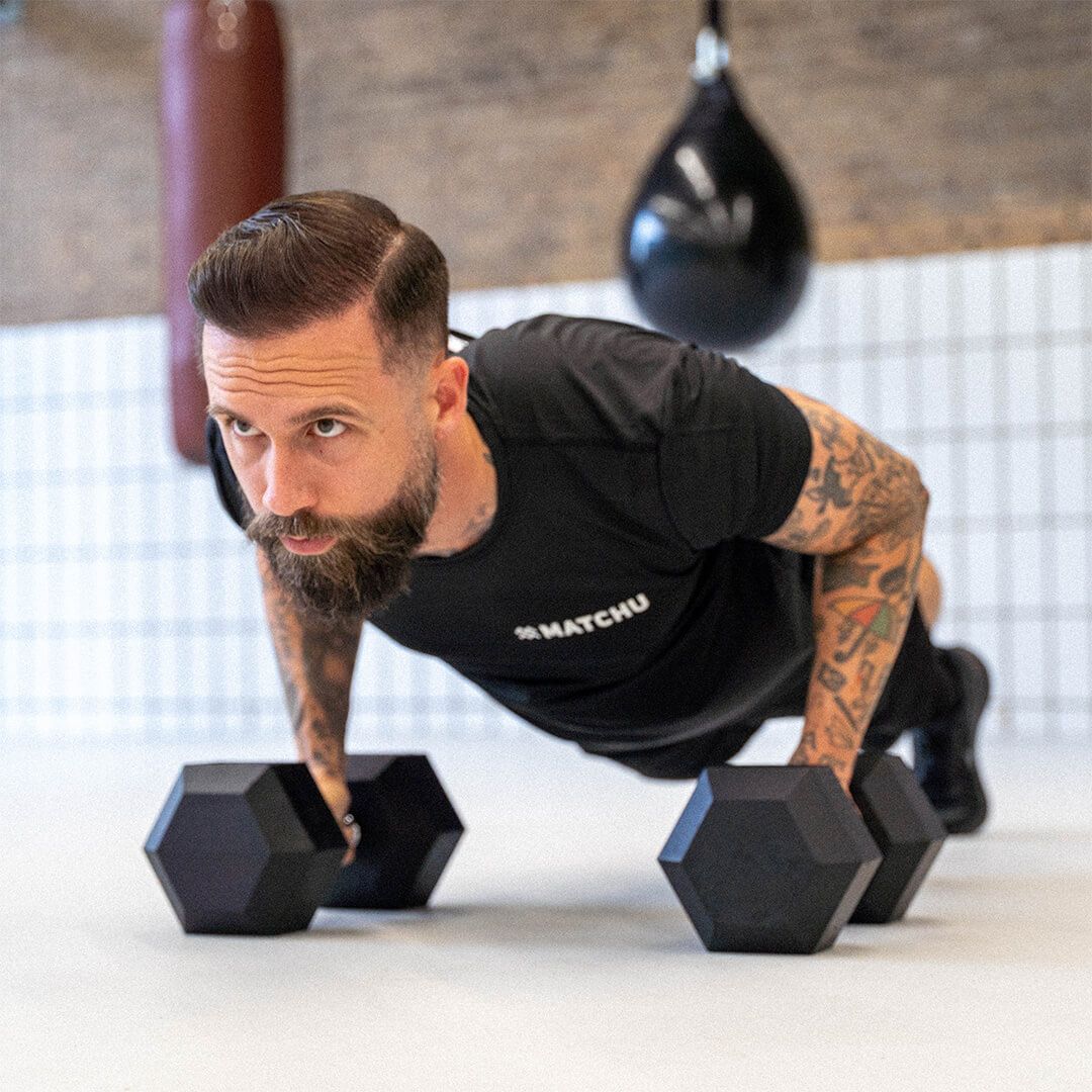 Mann macht Liegestütze mit zwei schwarzen Kurzhanteln. Er trägt ein schwarzes Shirt mit dem Logo "MATCHU".