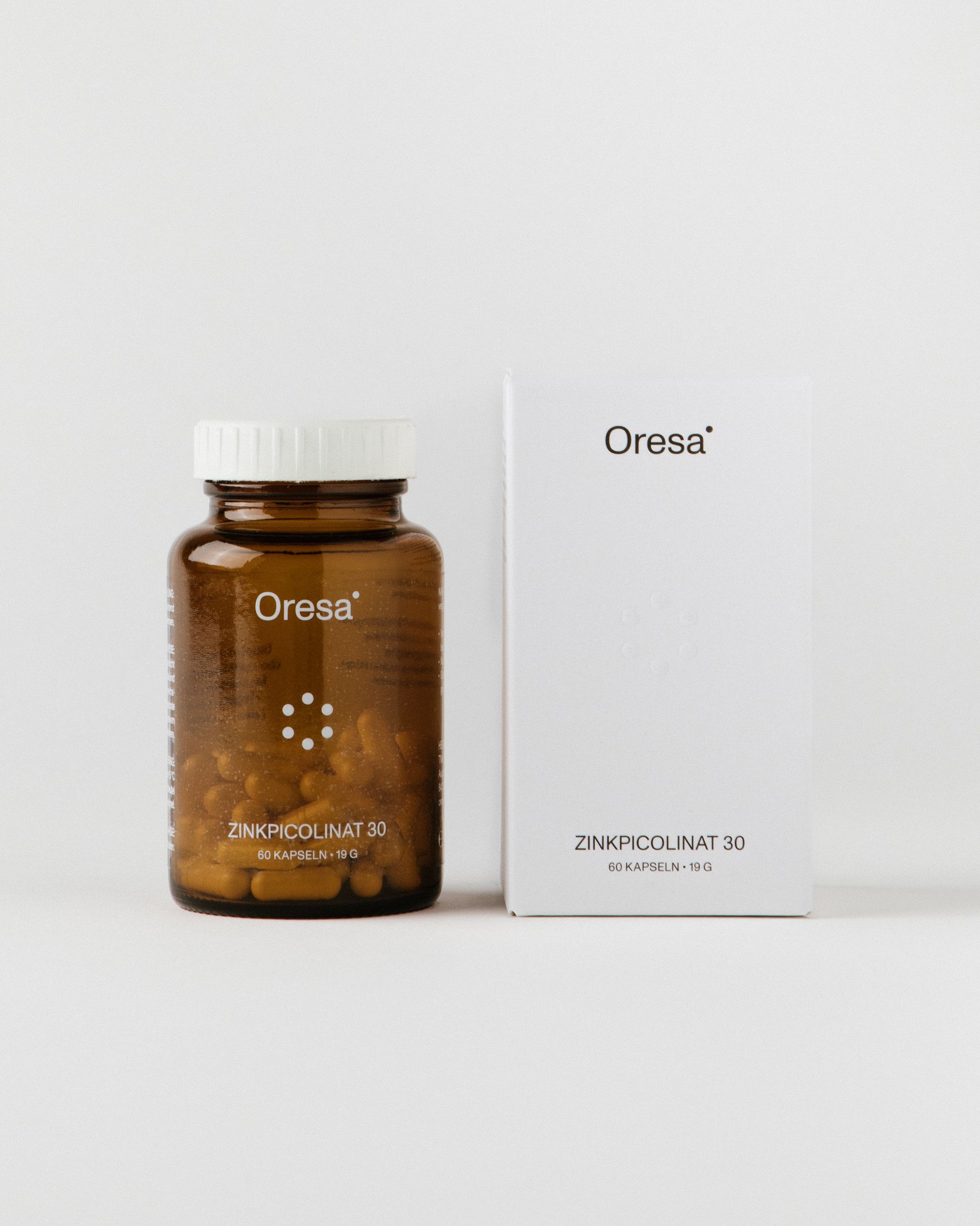 Braune Glasflasche und weiße Schachtel. Auf der Flasche und Schachtel steht "Oresa Zinkpicolinat 30". Kapseln sind in der Flasche sichtbar.