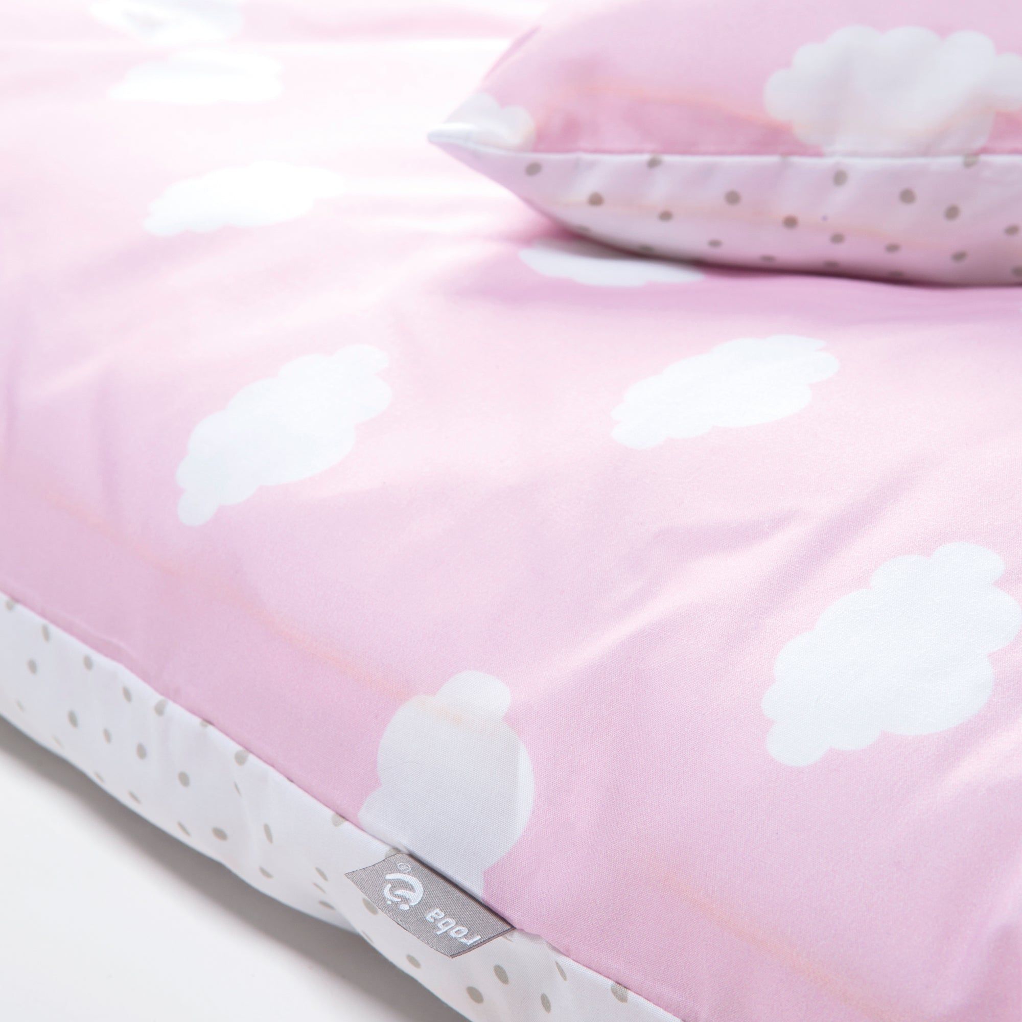 Detailansicht der Babybettwäsche. Rosa Stoff mit weißen Wolken und gepunktetem Rand. Label mit Logo.