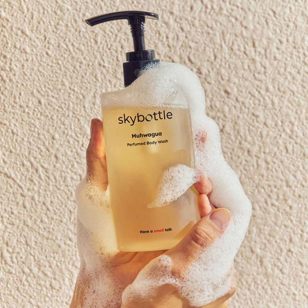 Hände halten eine Flasche SKYBOTTLE Muhwagua Duschgel, umgeben von Schaum. Aufschrift: Muhwagua, Perfumed Body Wash. Unter: Have a smell talk.
