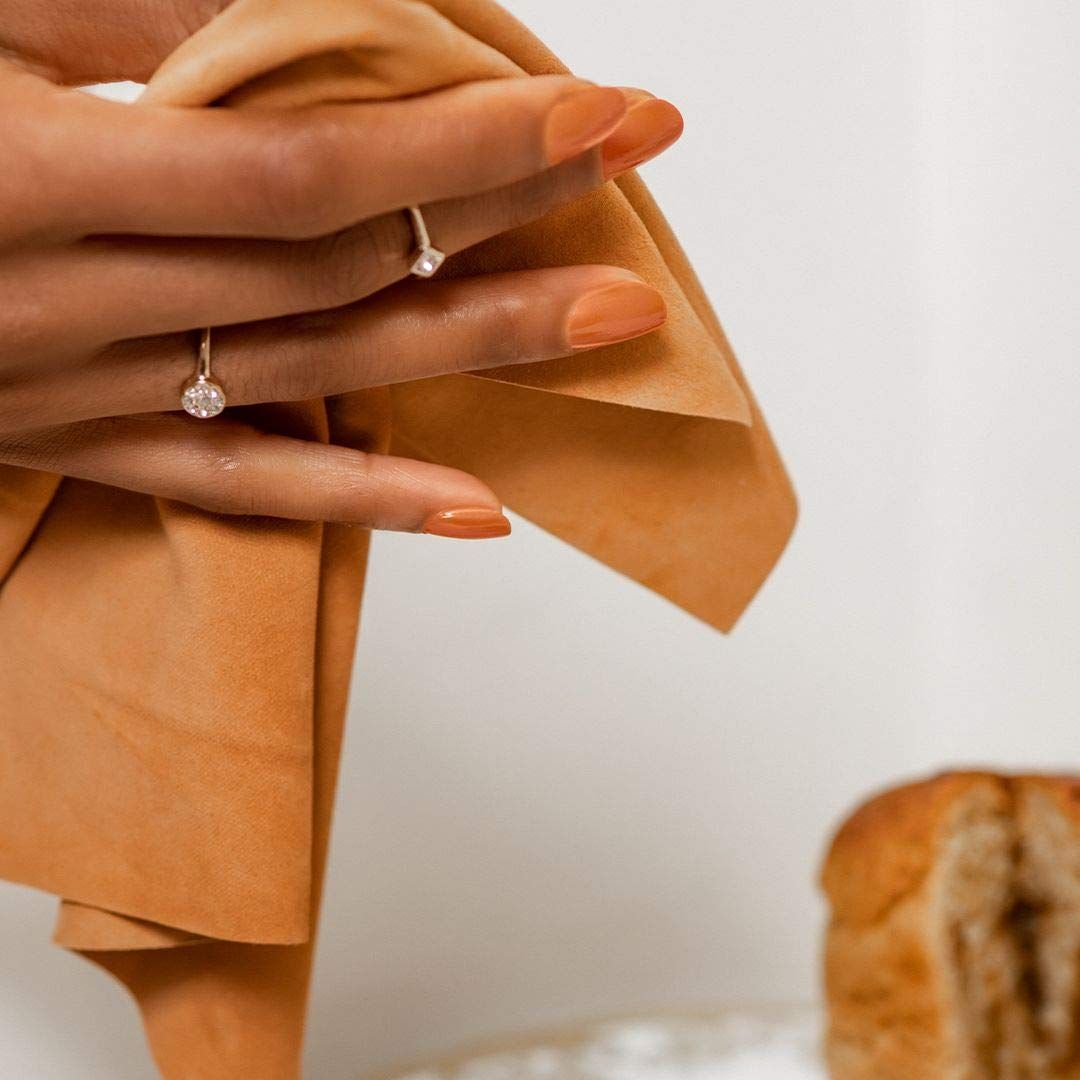 Hand mit orange lackierten Nägeln hält ein braunes Tuch. Zwei Ringe an den Fingern.