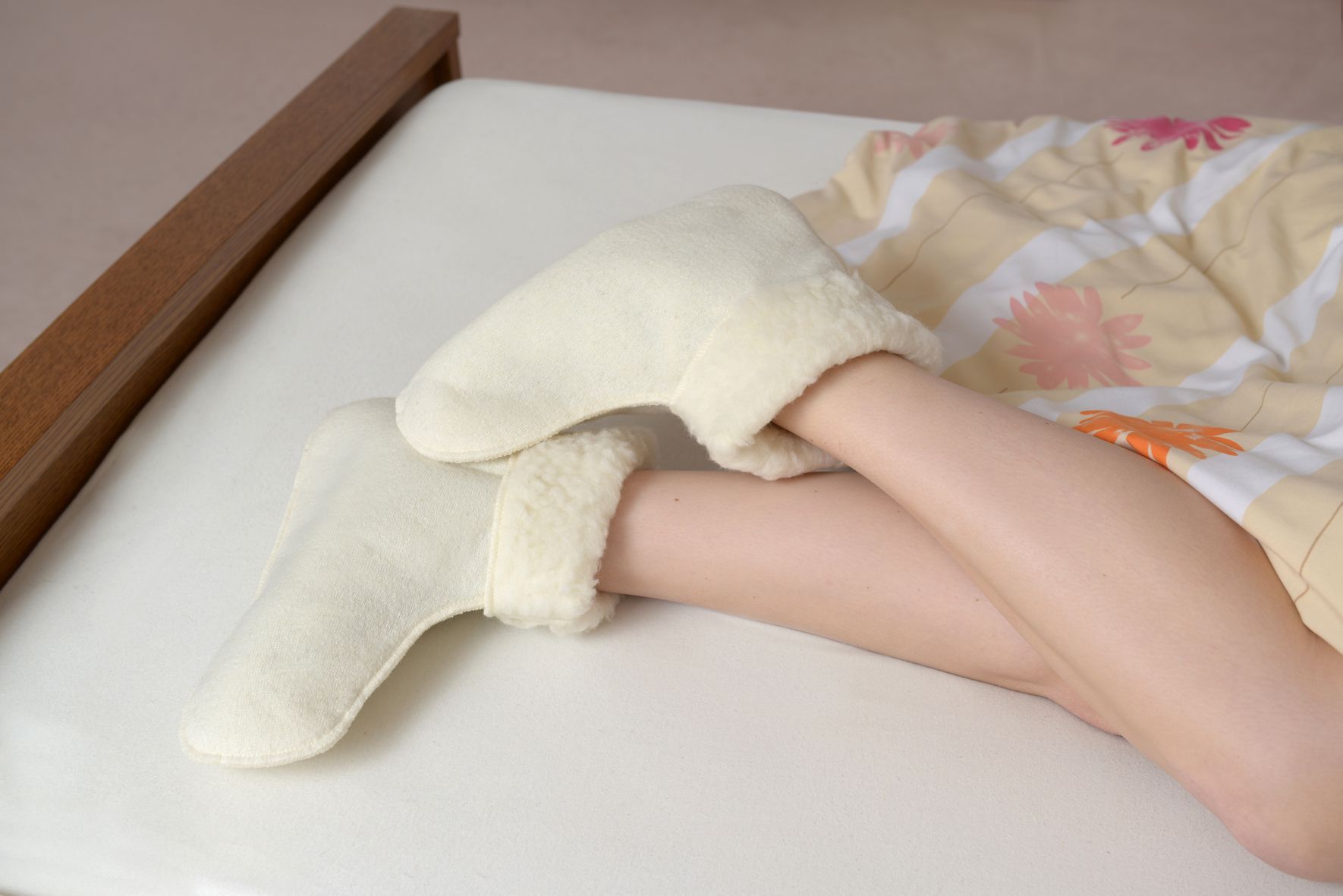 Person trägt cremefarbene Schurwoll-Bettschuhe. Füße und Unterschenkel auf Bett mit Decke.