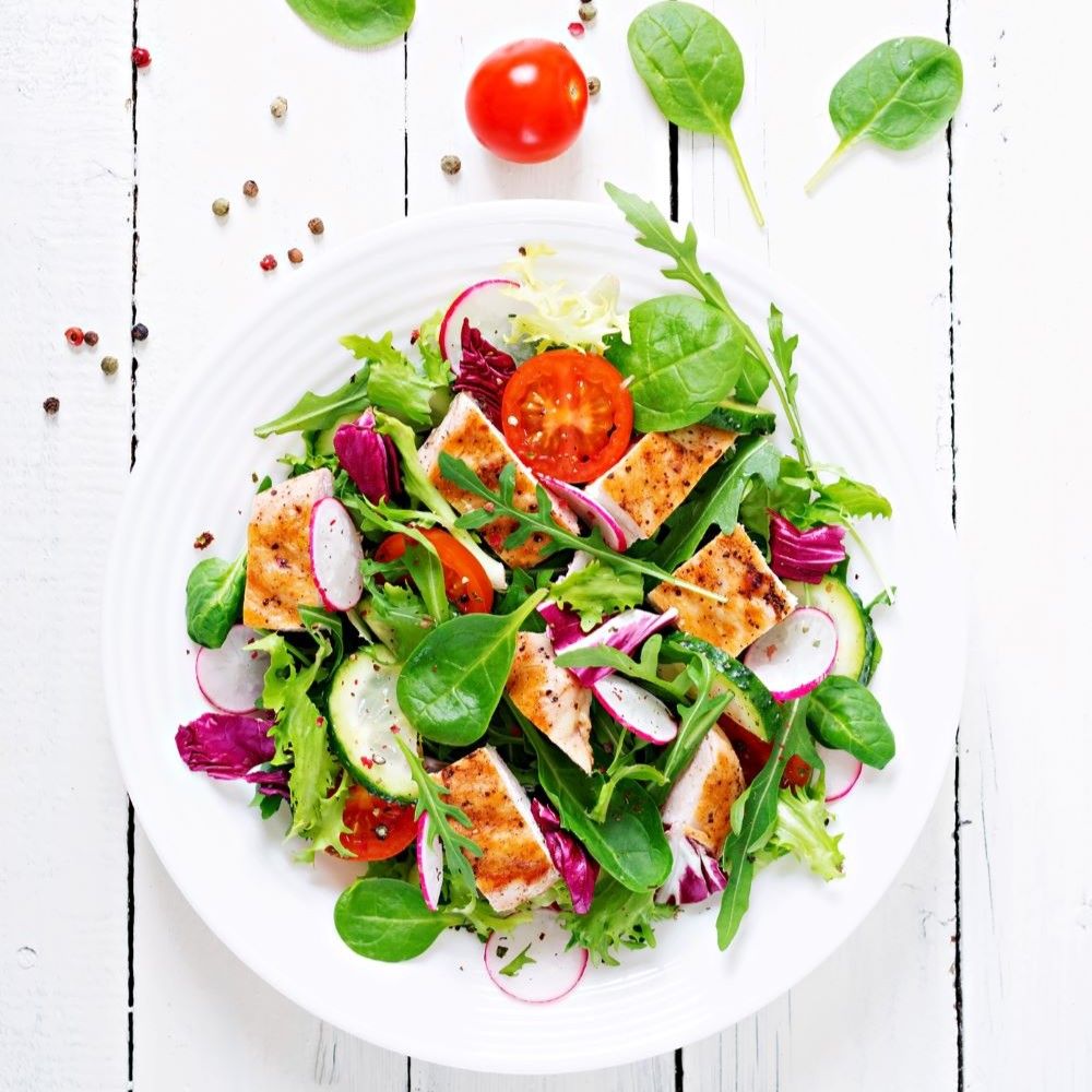 Salat mit Hähnchen, Tomaten, Radieschen und Blattsalat auf einem weißen Teller. Auf einem weißen Holztisch.