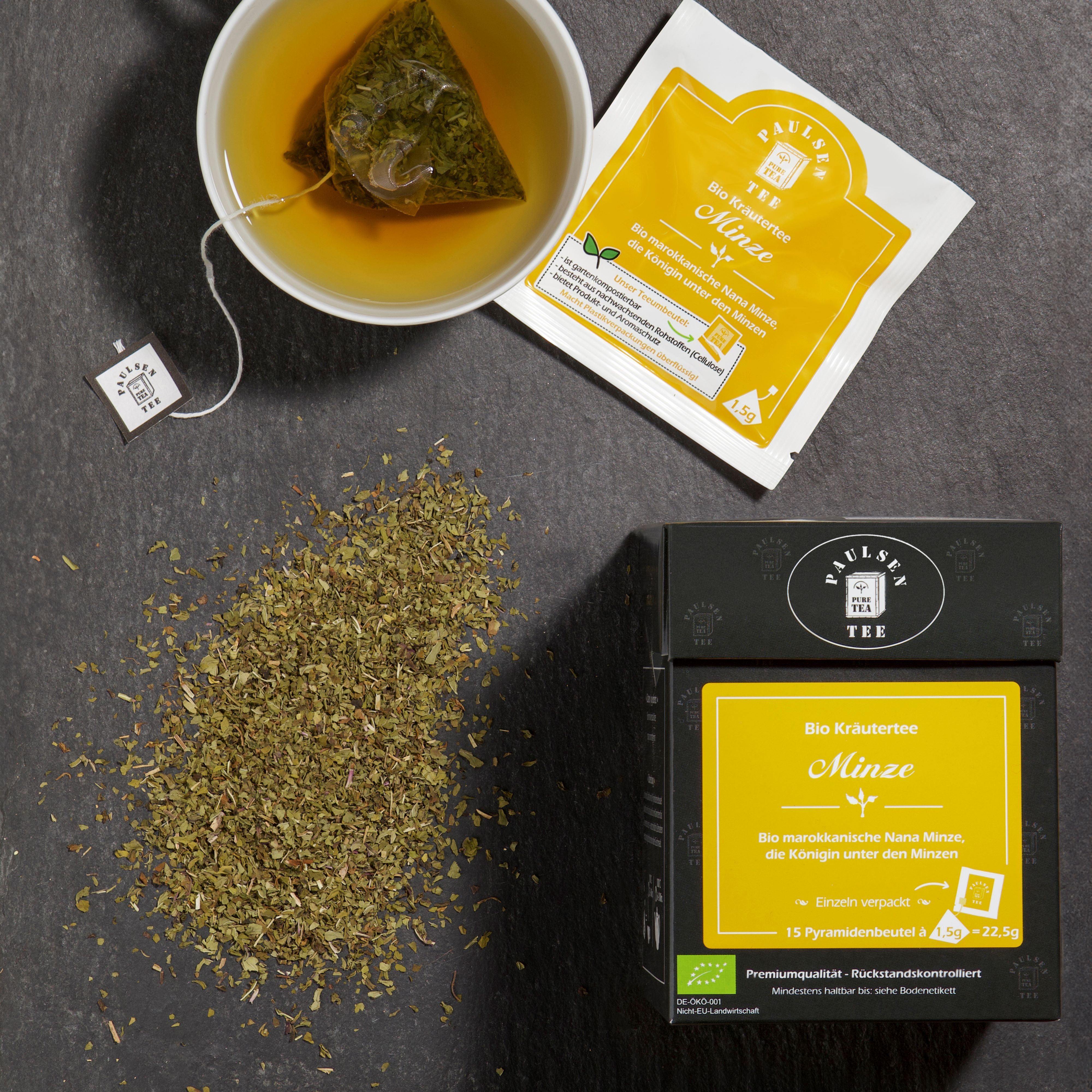 Tasse Tee, Teebeutel und loser Tee auf dunklem Untergrund. Box und Teebeutel mit Aufschrift: Bio Kräutertee Minze.