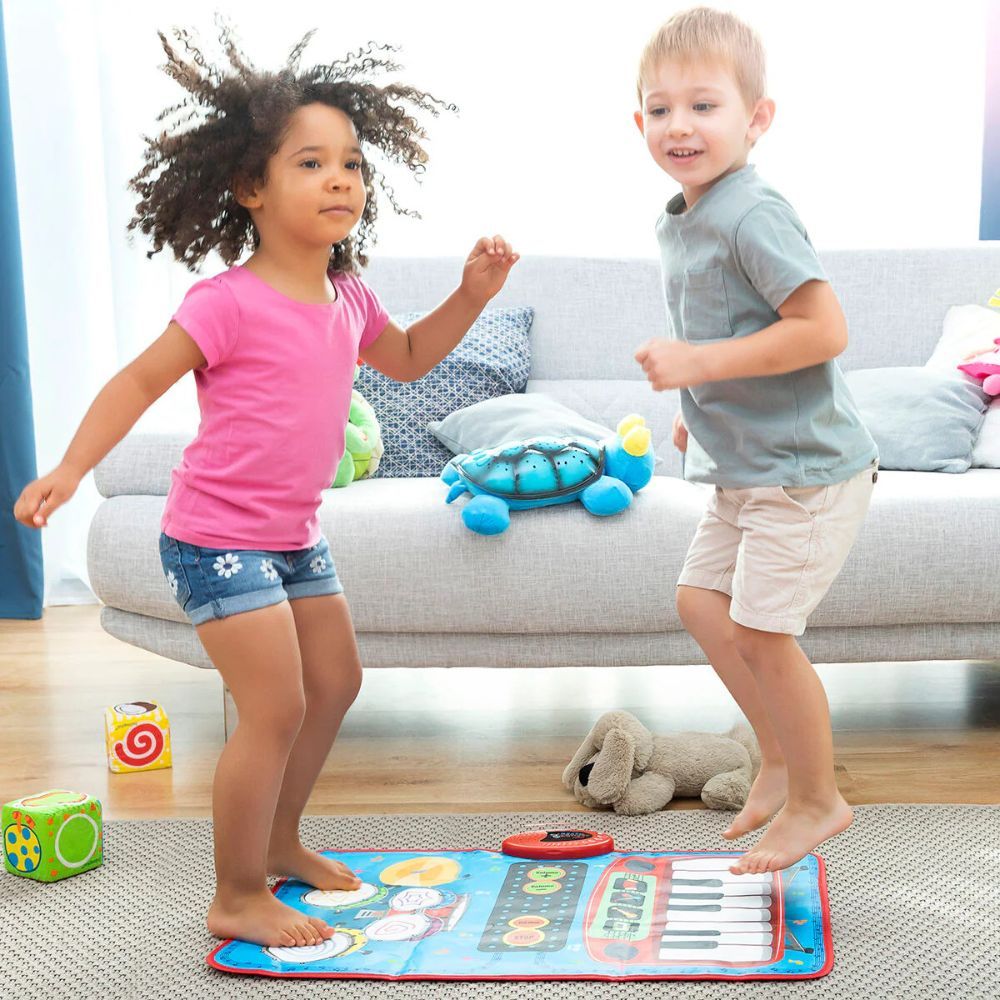 Zwei Kinder tanzen auf einer blauen Musikmatte. Die Matte hat Trommeln und Klaviertasten. Ein Sofa und Spielzeug sind im Hintergrund.