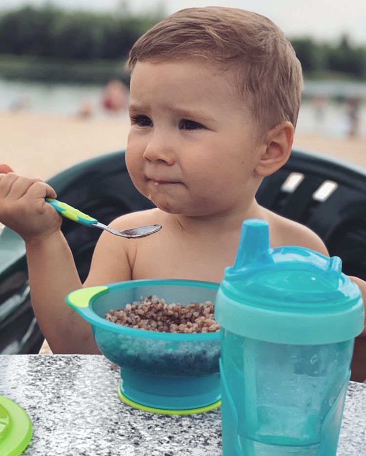 Kleinkind isst Brei und hat einen blauen Trinkbecher mit Strohhalm neben sich.