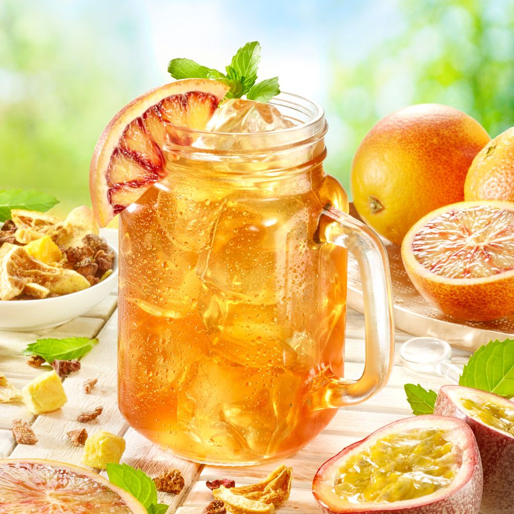 Glas mit Eistee, Orangenscheibe, Minze und Eiswürfeln. Daneben Orangen und Maracujas.