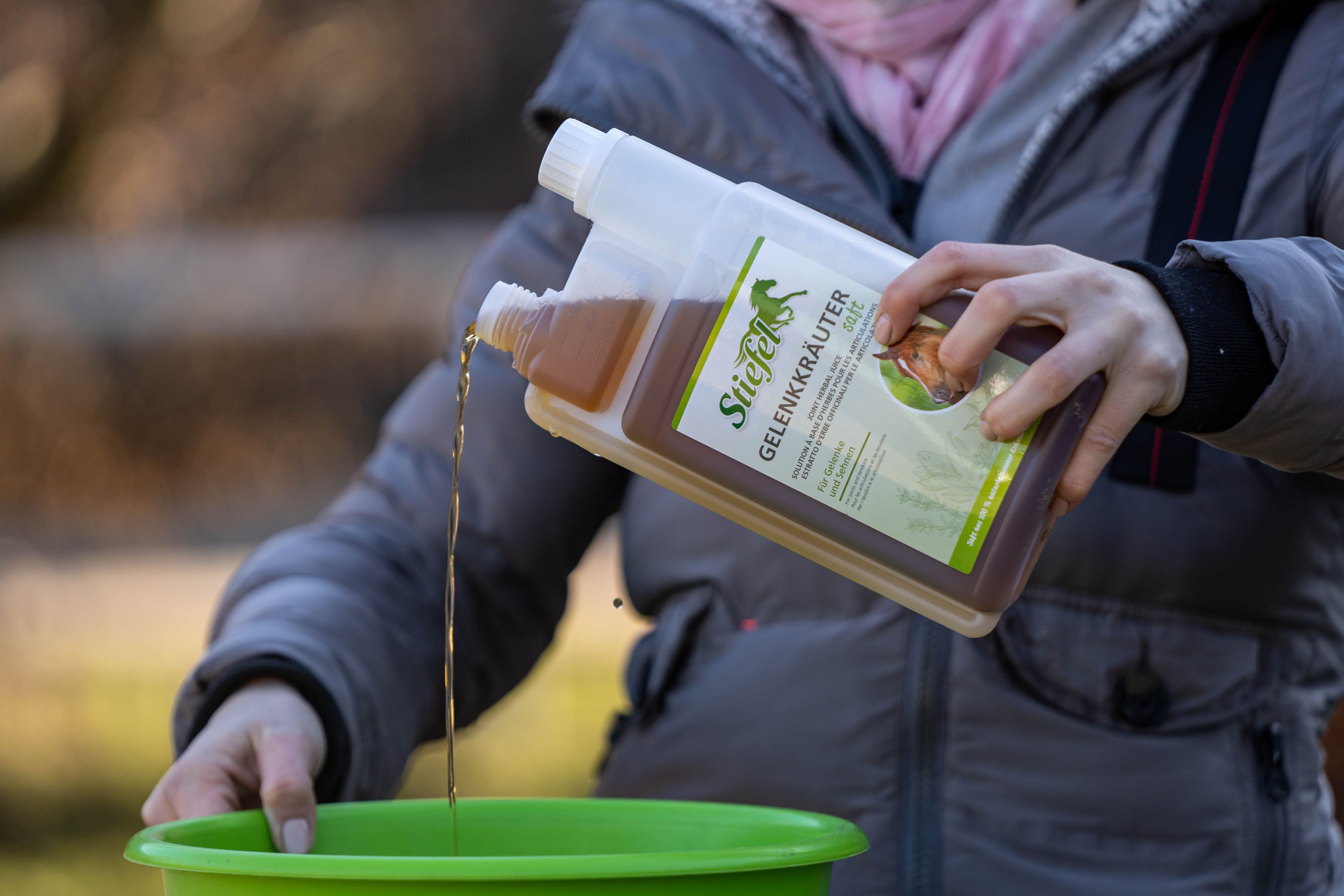 Person gießt Stiefel Gelenkkräuter Saft in einen grünen Behälter. Die Flasche ist transparent und enthält eine braune Flüssigkeit. Aufschrift: Für Gelenke und Sehnen.