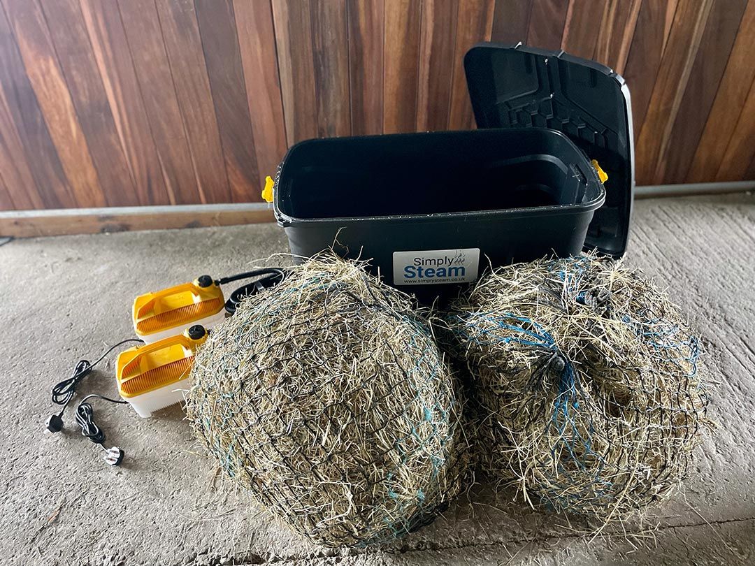 Schwarzer Behälter mit geöffnetem Deckel, Simply Steam-Logo. Zwei gelb-weiße Geräte mit Kabeln. Zwei Heunetze daneben. Holzverkleidung im Hintergrund.