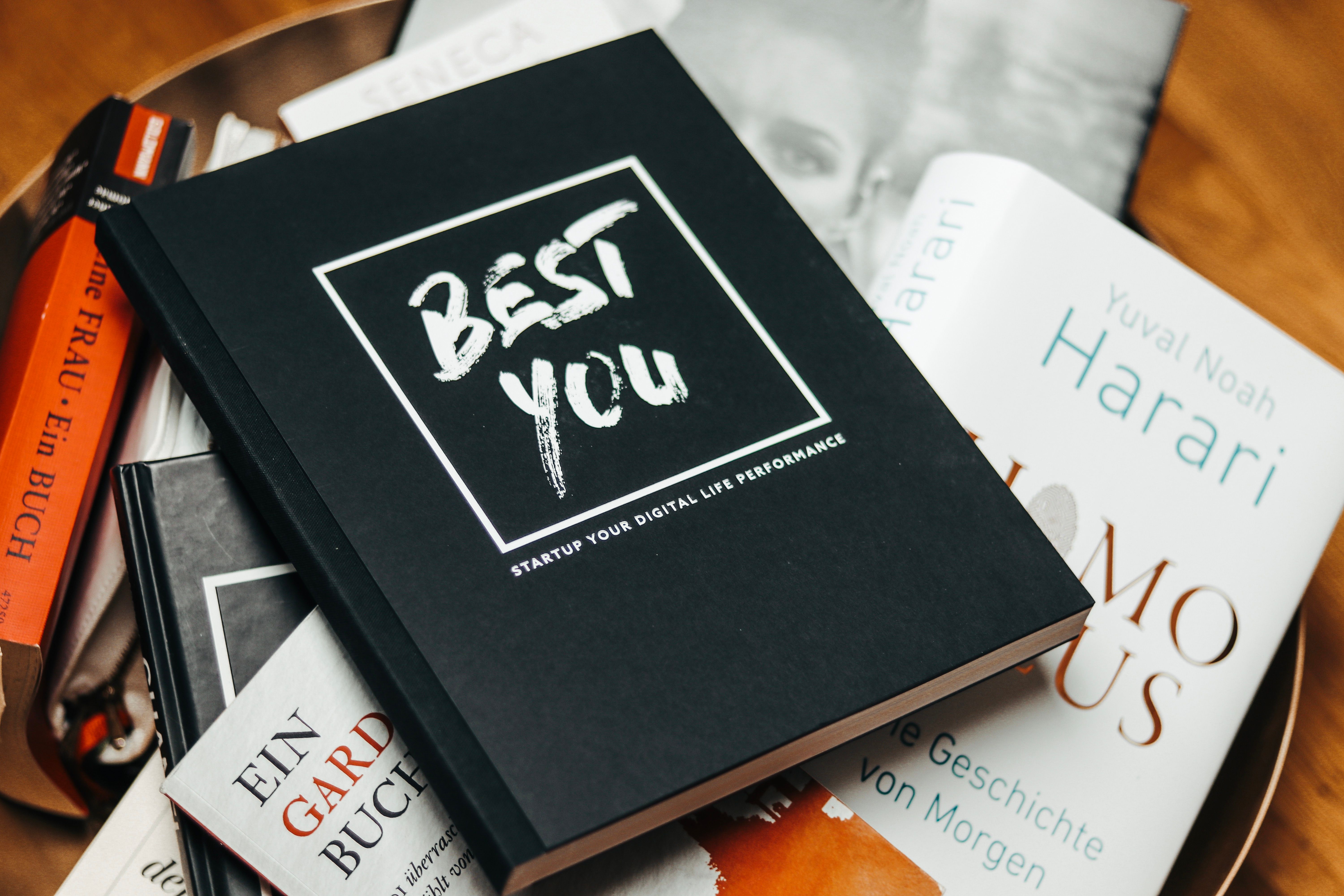 Schwarzes Buch mit weißem Schriftzug „BEST YOU“. Darunter „STARTUP YOUR DIGITAL LIFE PERFORMANCE“. Auf weiteren Büchern liegend.