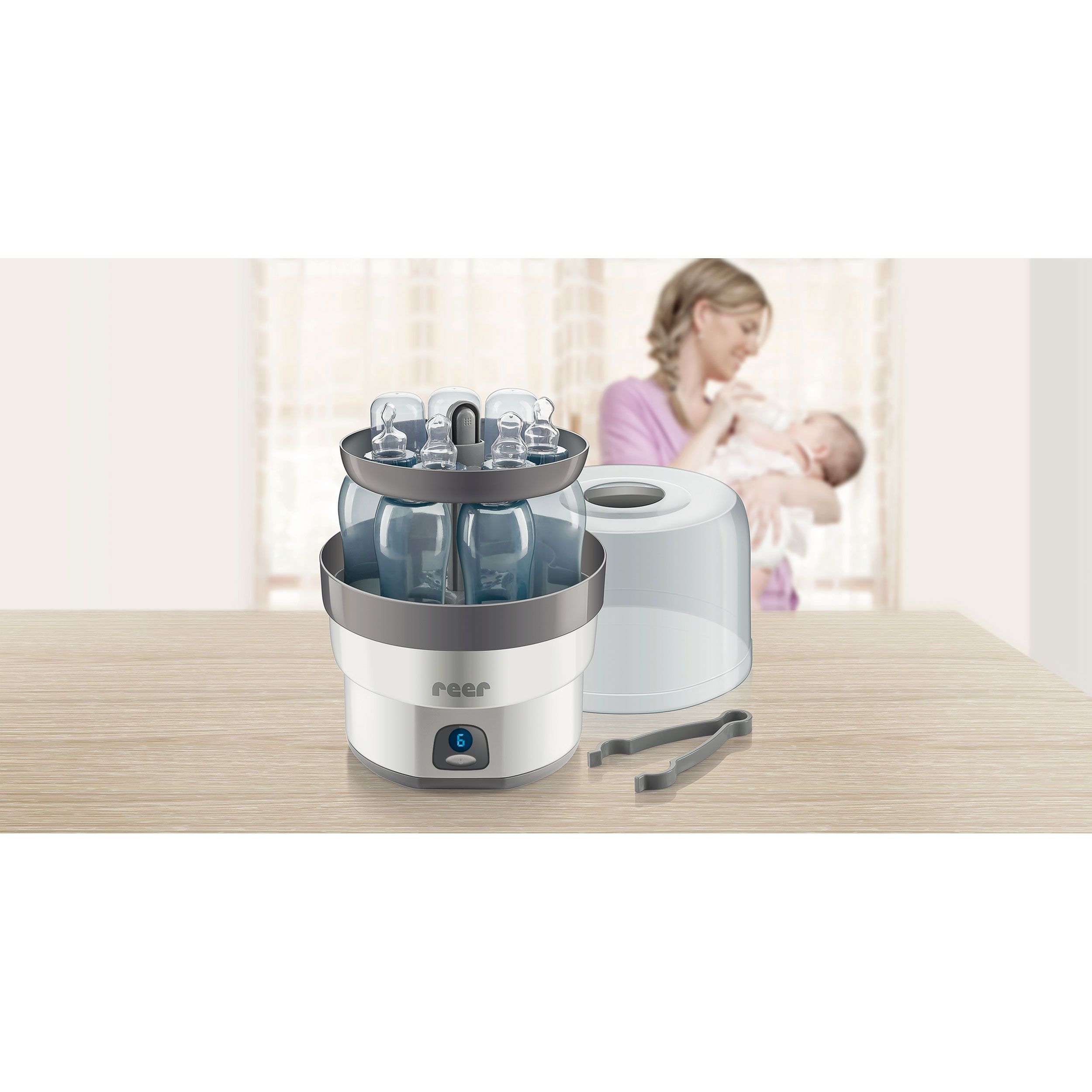 Sterilisator mit Flaschen. Graue Zange. Marke: reer. Frau mit Baby im Hintergrund.