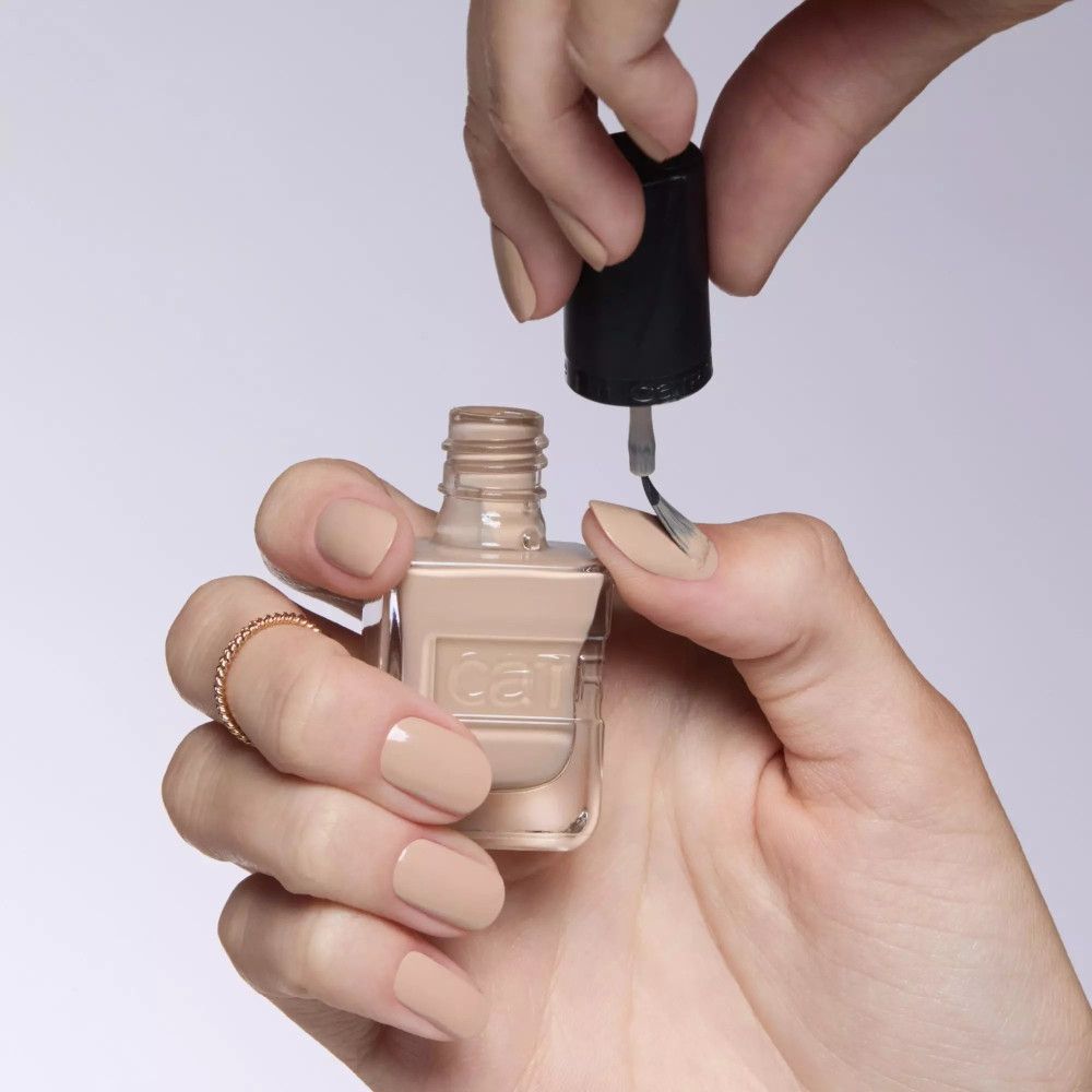 Hände mit Nagellackflasche und Pinsel. Beige Nagellackfarbe. Schwarzer Deckel mit "Catrice"-Schriftzug. Lackierte Nägel.