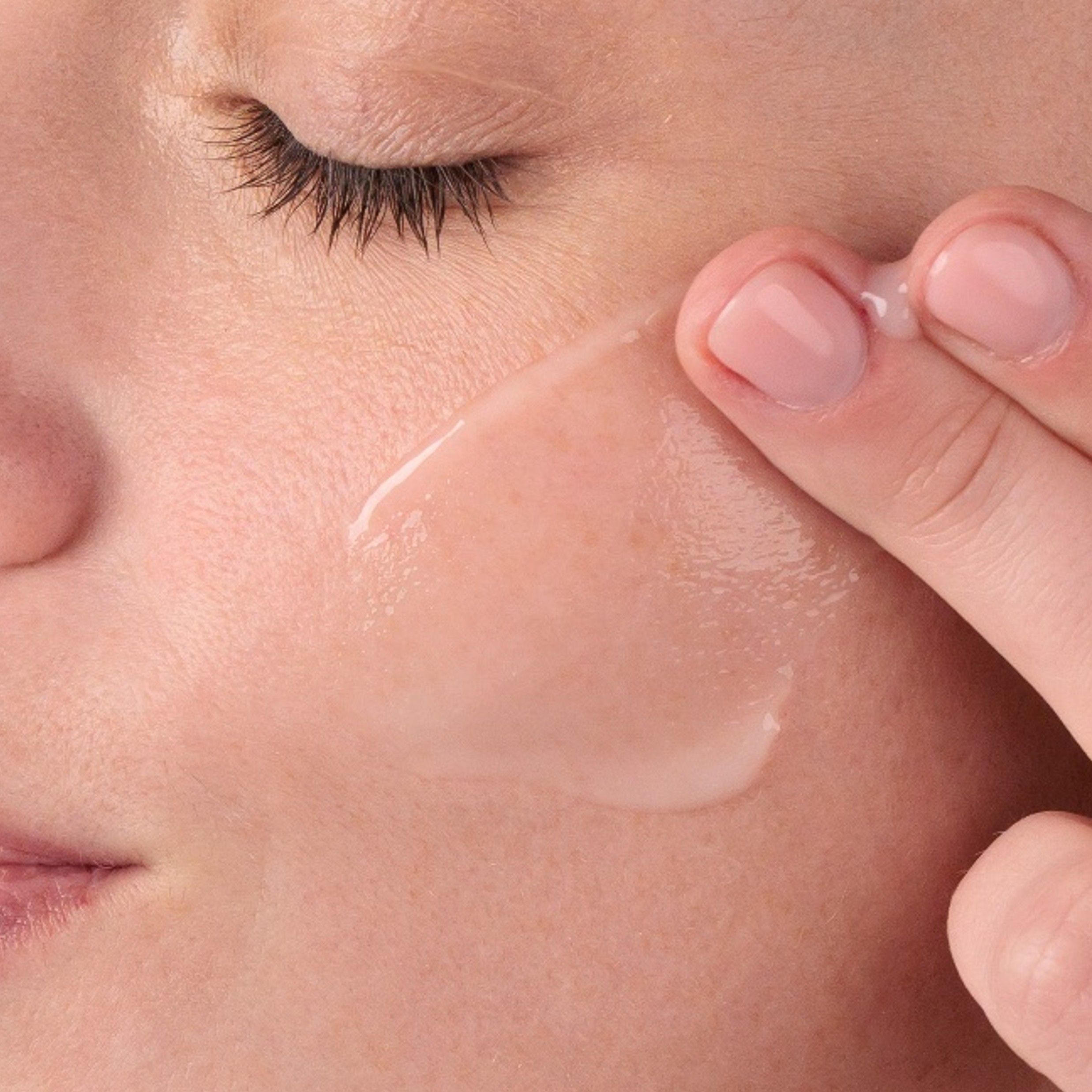 Nahaufnahme: Finger tragen Serum auf Gesicht auf. Klare, transparente Flüssigkeit. Haut wirkt feucht.