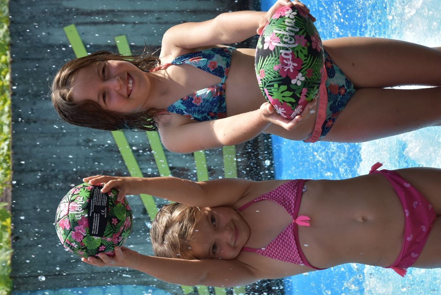 Zwei Mädchen im Wasser spielen mit Bällen mit tropischem Blumenmuster. Eine wirft, eine hält.