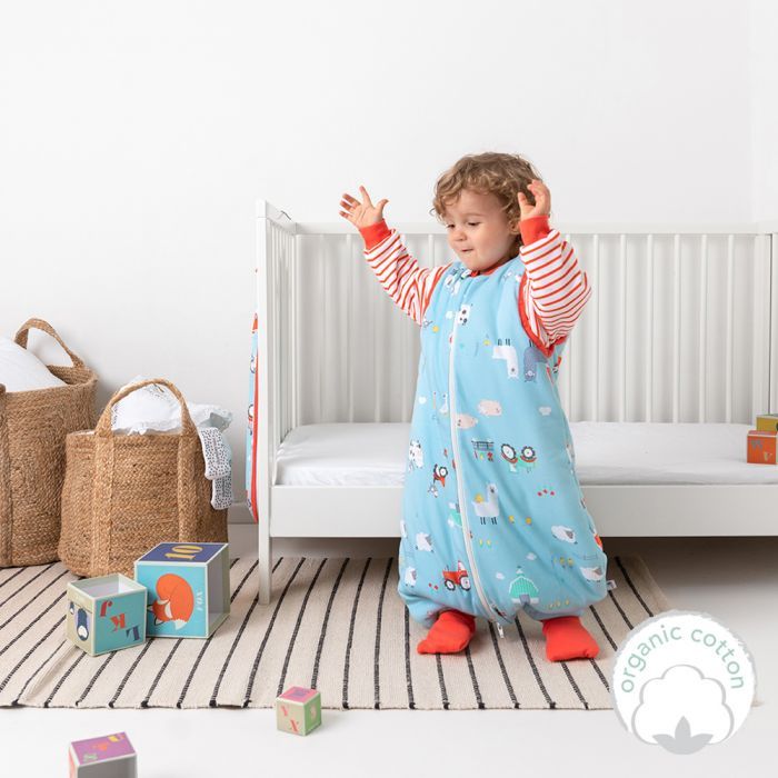 Kleinkind in blauem Schlafsack mit Bauernhof-Motiv, rote Füße. Steht vor einem Bett, umgeben von Spielzeug.