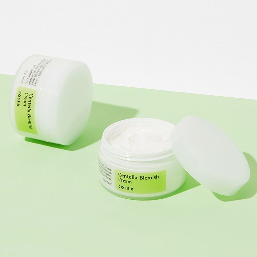 Zwei COSRX Centella Blemish Cream Tiegel, einer offen, auf grünem Untergrund.