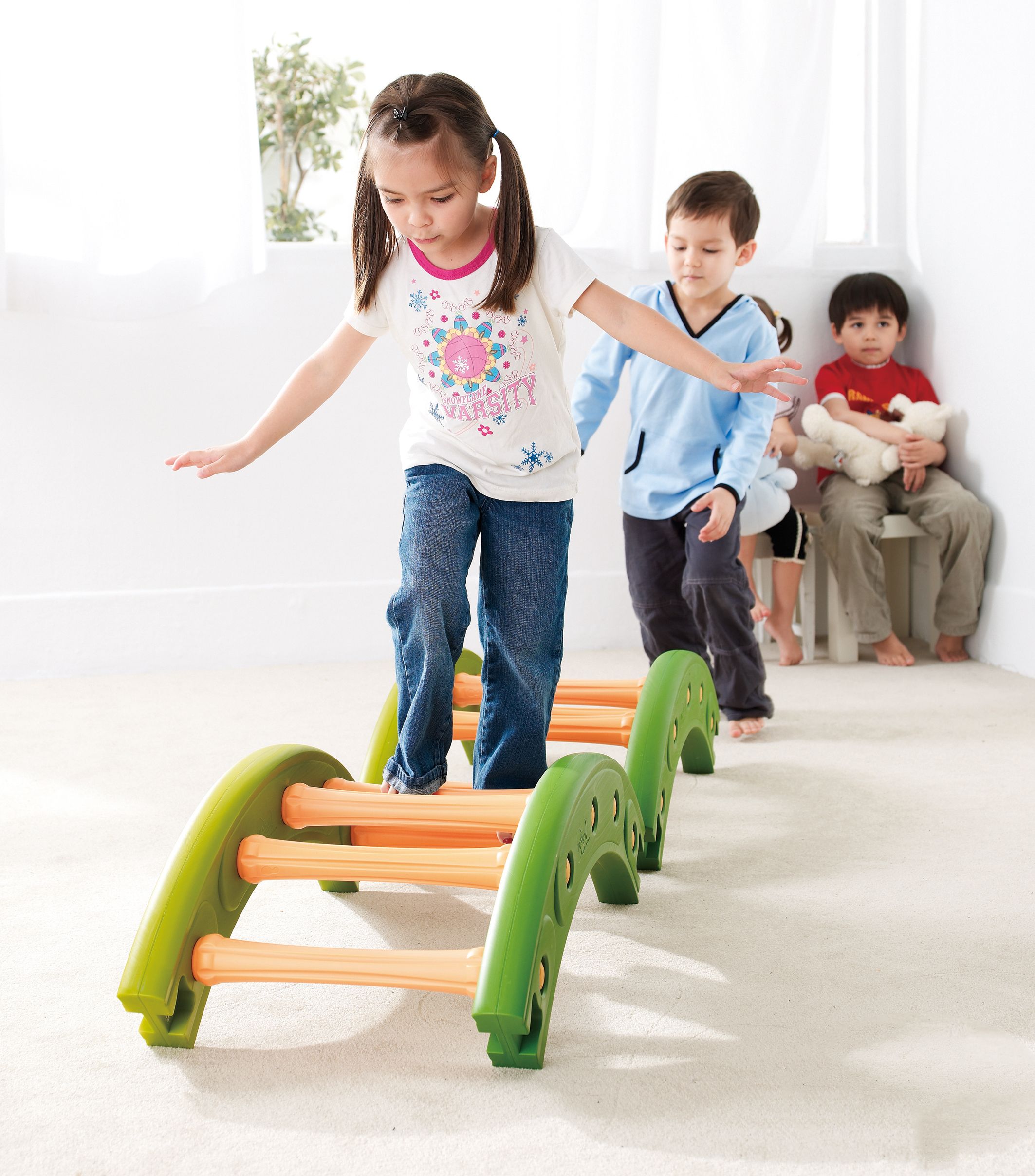 Grüne Balance-Brücke mit orangefarbenen Stützen. Ein Kind balanciert darauf, weitere Kinder folgen.