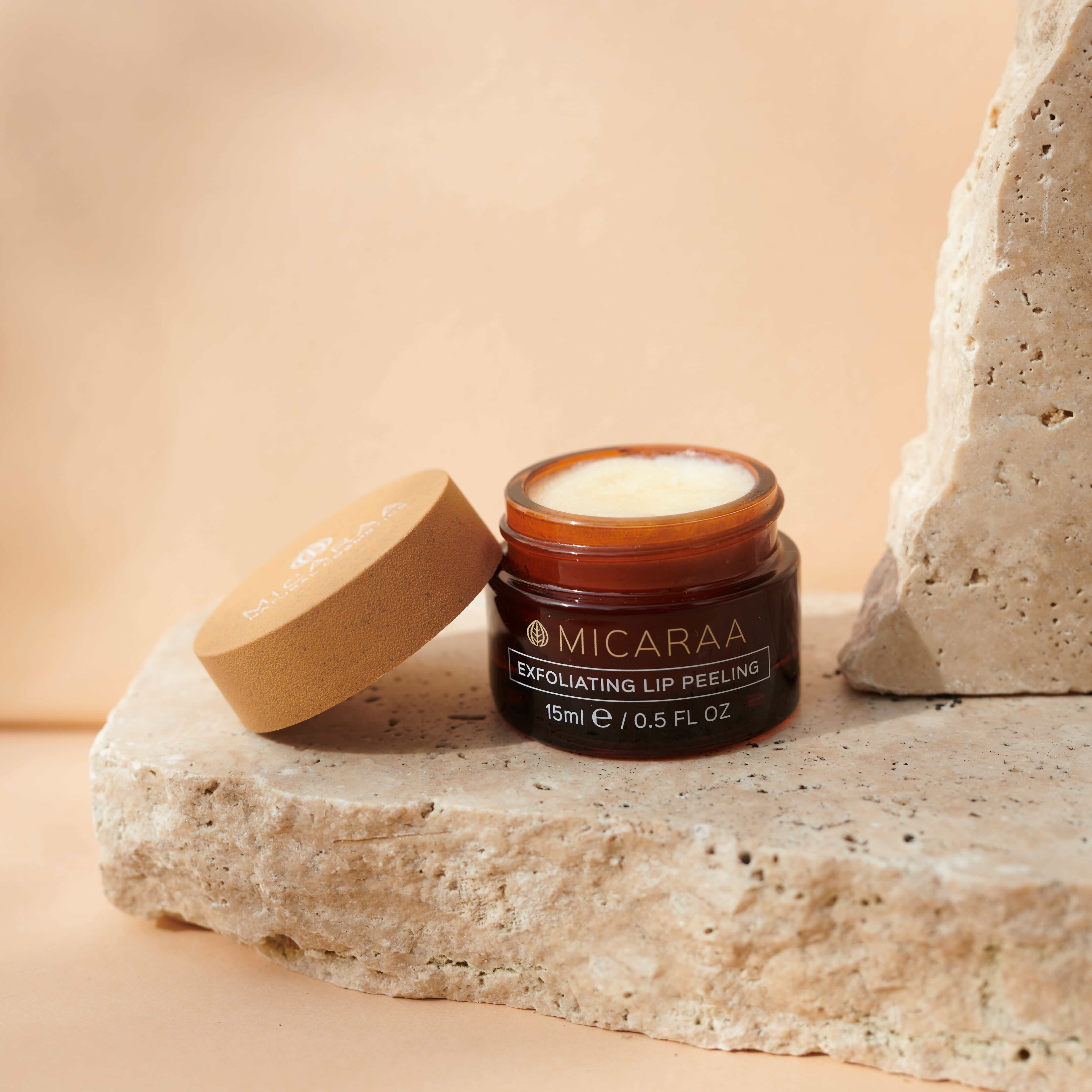Offene Dose mit Peeling. Deckel daneben. Aufschrift: MICARAA, Exfoliating Lip Peeling, 15ml / 0.5 FL OZ. Auf Stein.