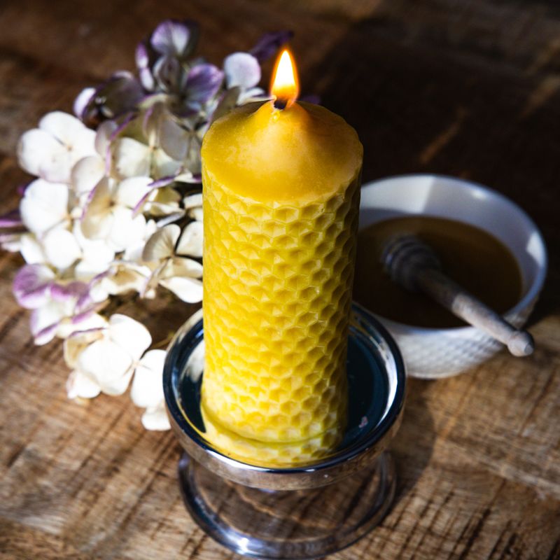Gelbe Wachskerze mit Wabenmuster, brennend. Auf einem silbernen Kerzenhalter, neben einer Schale mit Honig und Blumen.