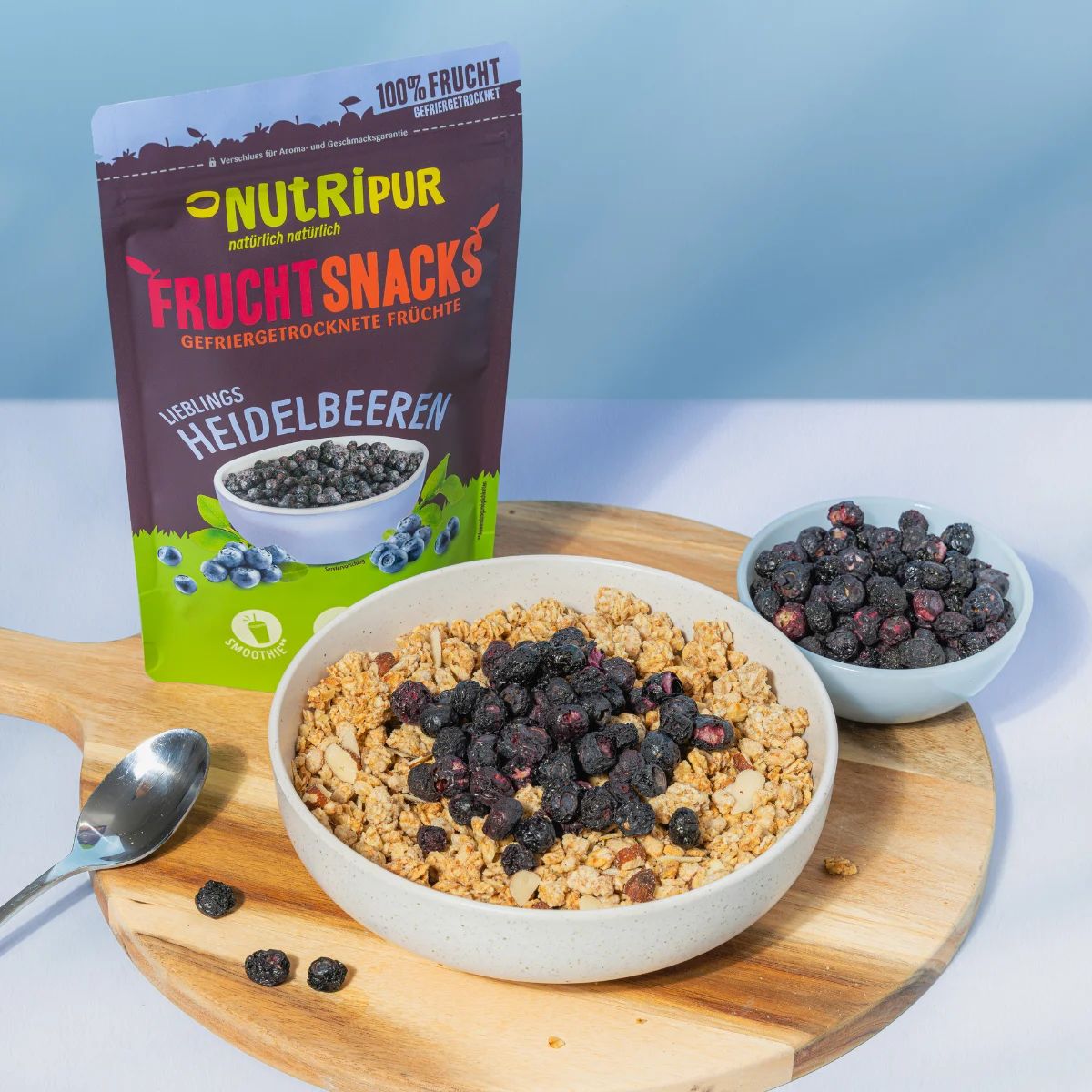 Beutel und Schalen mit gefriergetrockneten Heidelbeeren. Müsli mit Beeren in Schale. Auf Holzbrett mit Löffel.