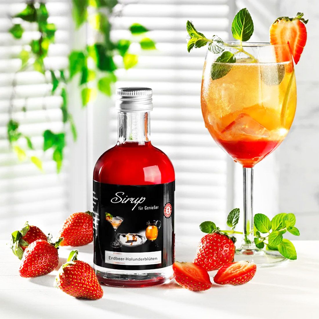 Glasflasche mit roter Flüssigkeit neben einem Glascocktail. Erdbeeren und Minze auf weißem Untergrund.
