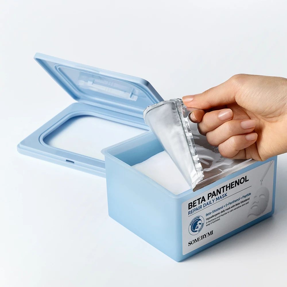 Hellblaue Box mit Deckel, gefüllt mit Masken. Eine Hand öffnet eine Maskenverpackung. Auf der Box steht "Beta Panthenol Repair Daily Mask".