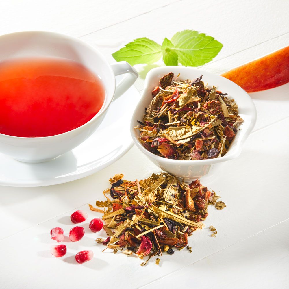 Tasse Tee, Schale mit Kräutern und Granatapfelkerne auf weißem Untergrund. Rote Flüssigkeit im Teebecher.