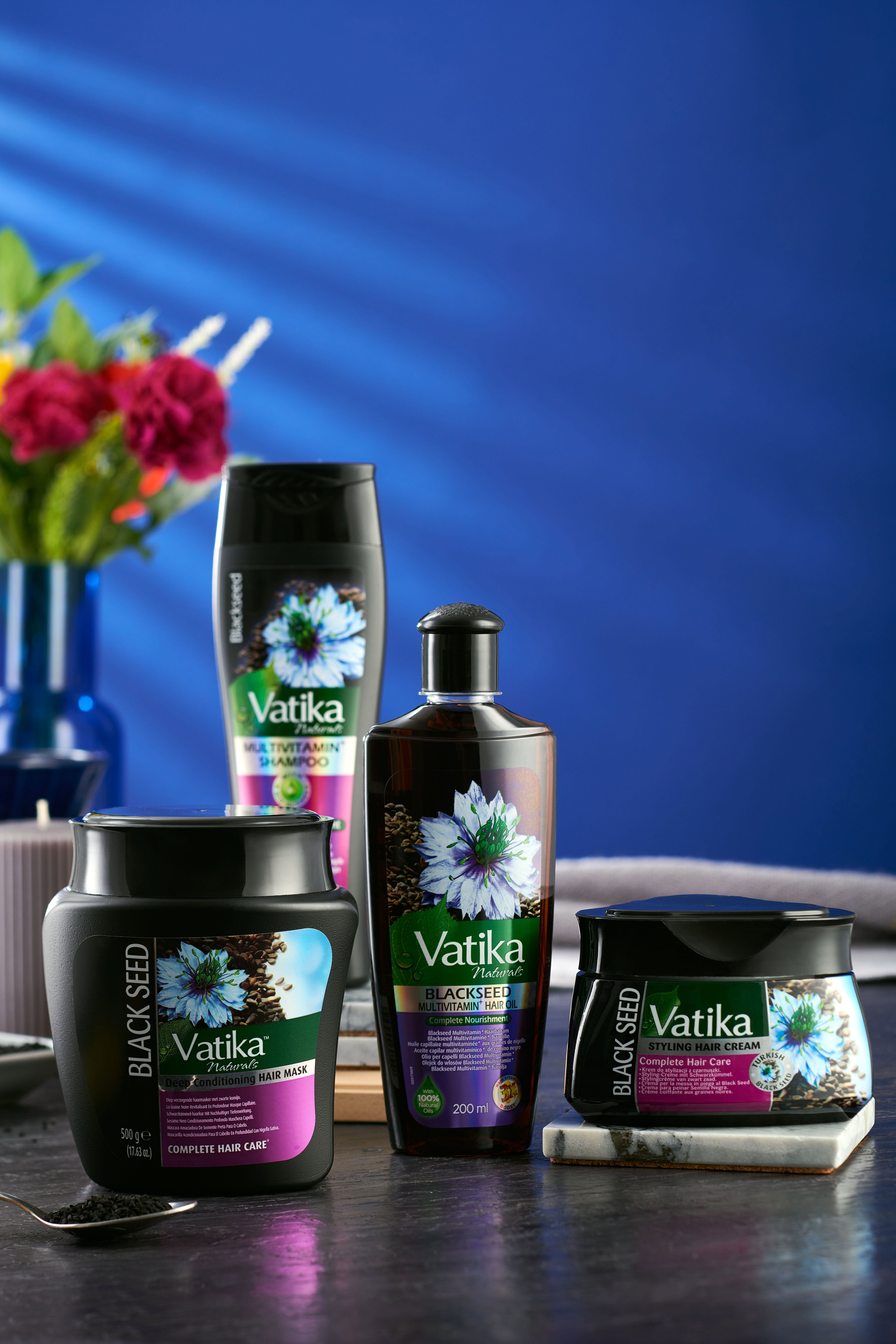 Produkt-Ensemble: Haarmaske, Shampoo, Öl und Creme. Schwarze Verpackungen mit Vatika-Logo und Schwarzkümmel-Abbildung. Auf Tisch platziert.